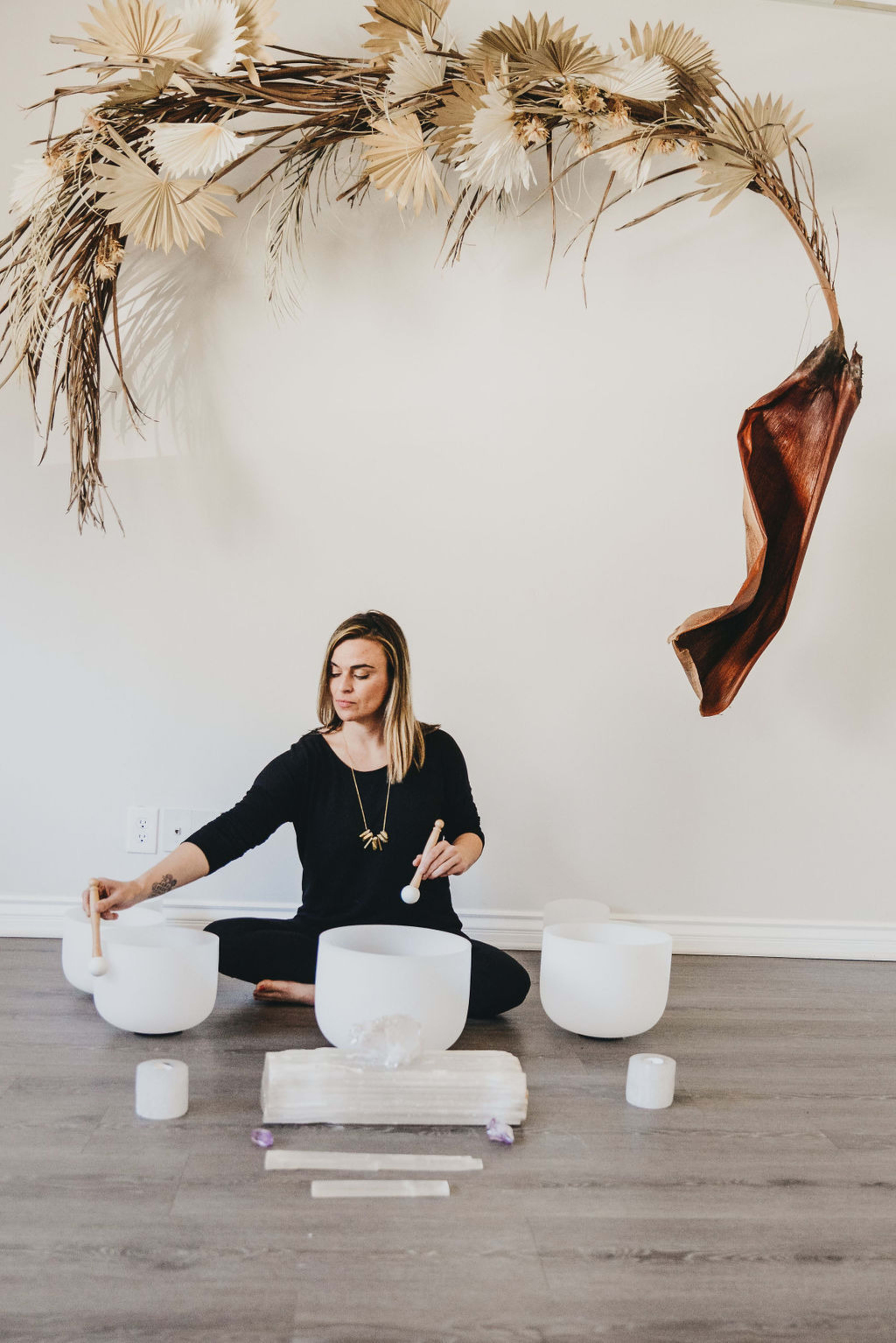 A woman sits on the floor, playing crystal singing bowls, with decorated wall elements made of dried leaves above her.