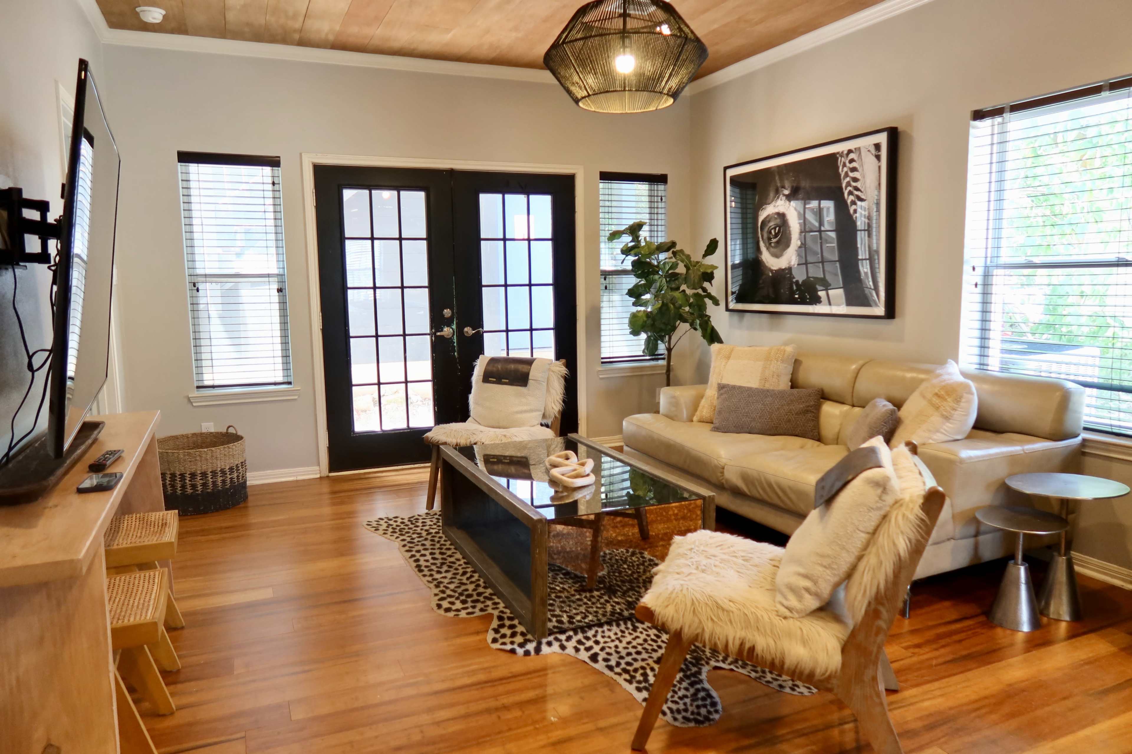 The image shows a modern living room featuring a light-colored sofa, a glass coffee table on a patterned rug, and large windows that allow natural light to fill the space.