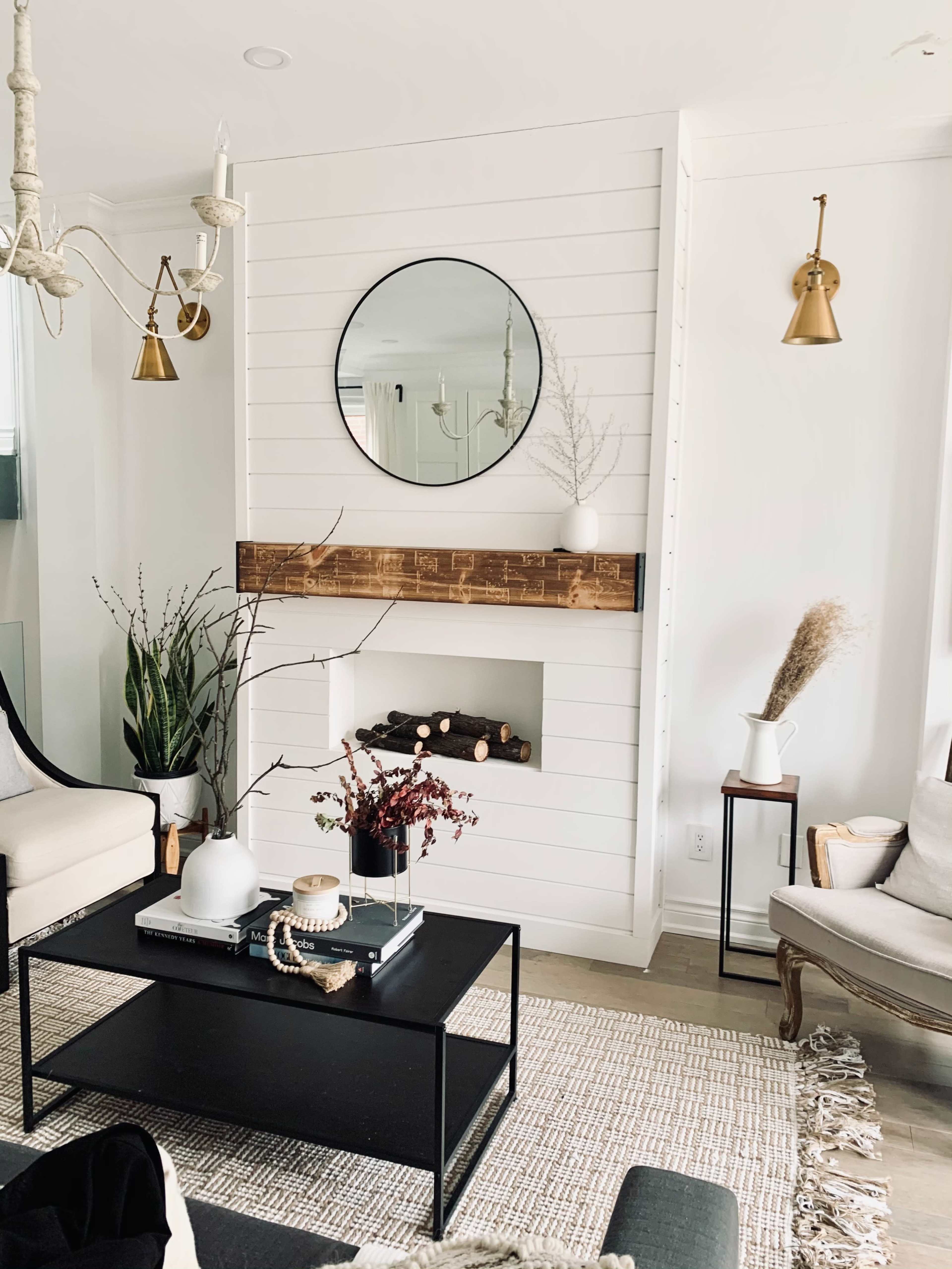 A modern living room with a white shiplap wall featuring a round mirror above a wooden mantel, a simple fireplace, and two seating areas, all decorated with plants and minimalistic decor.