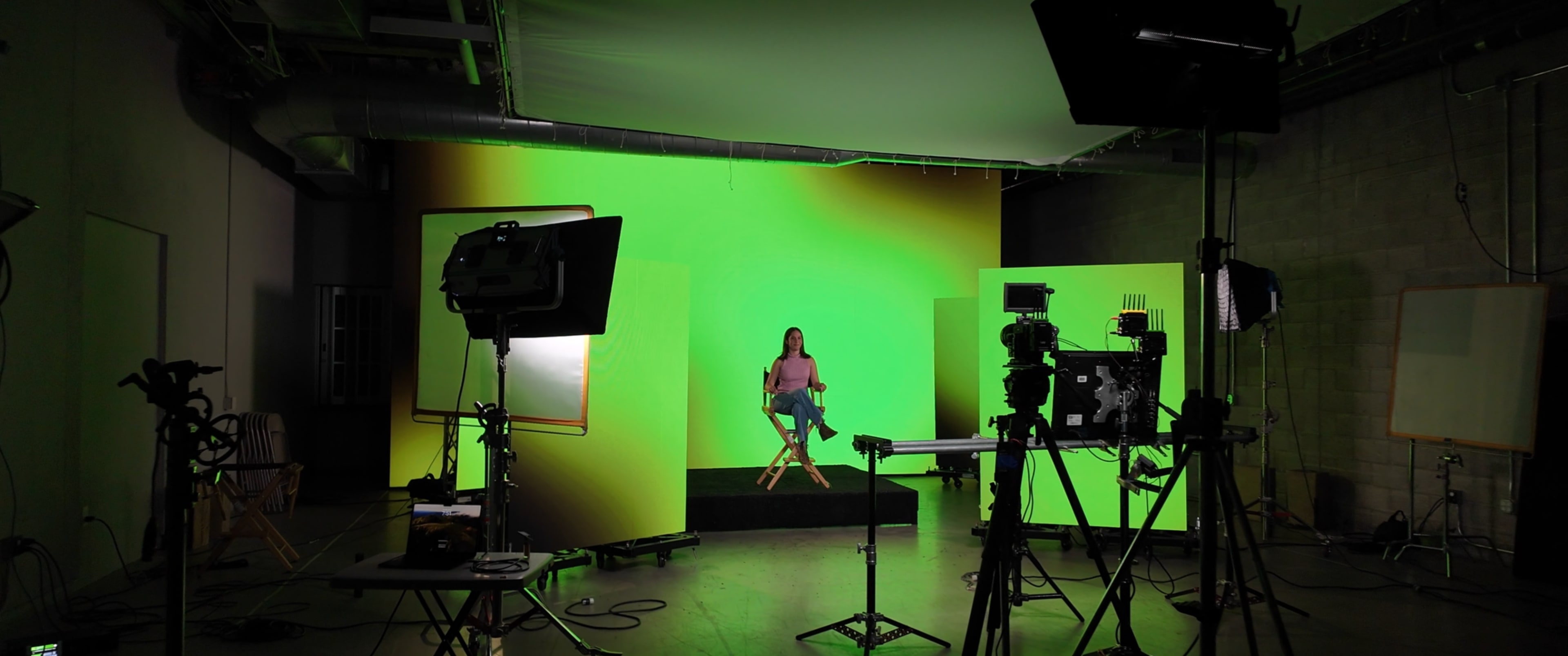 A person sits on a stool in front of a green screen in a well-equipped studio, surrounded by cameras and lighting equipment.