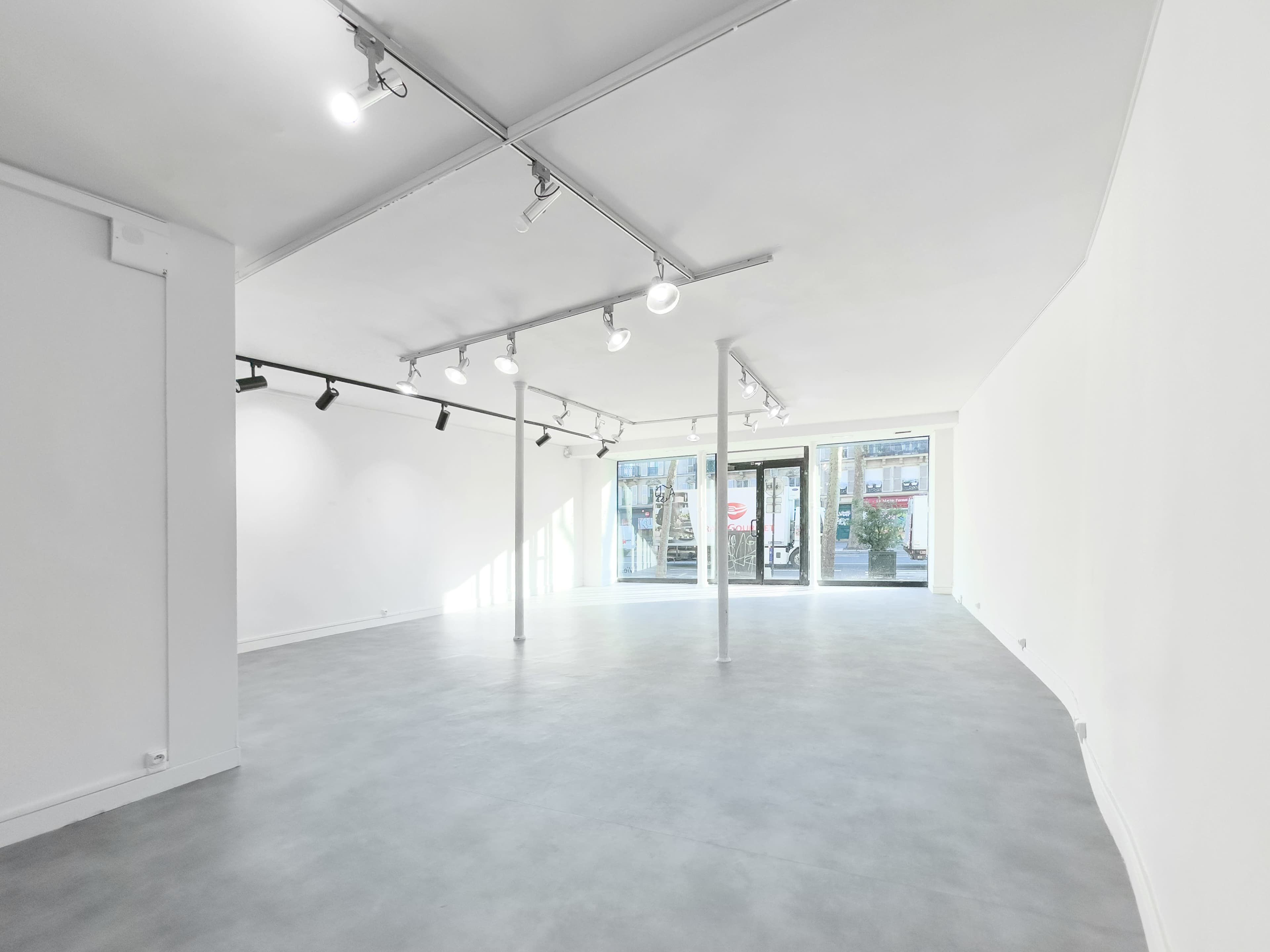 The image shows a spacious, empty room with a gray floor and white walls, featuring track lighting and large glass doors at one end.