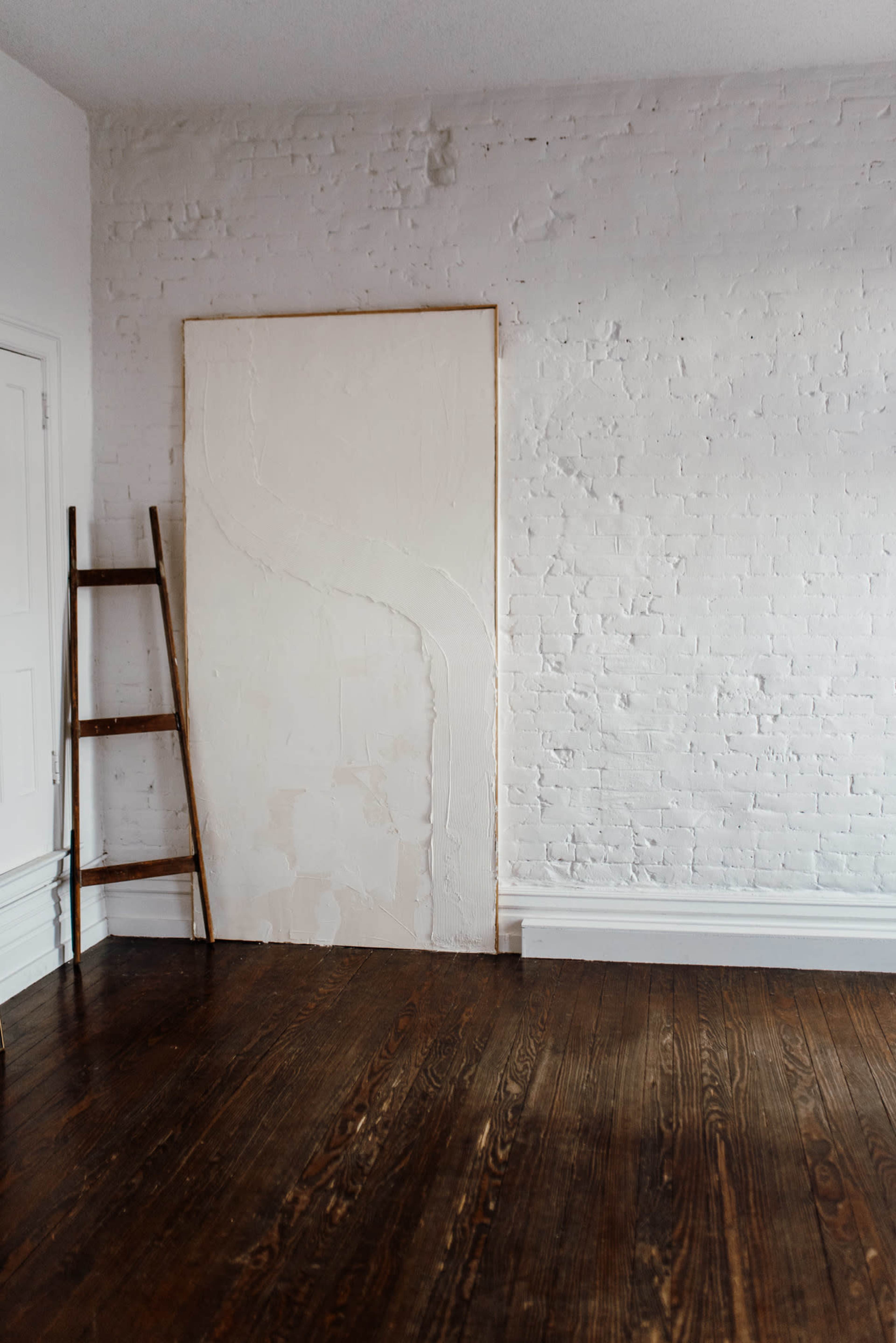 A minimalistic room with a white brick wall, a plain white panel, and a wooden ladder leaning against the wall, all set on dark wooden flooring.