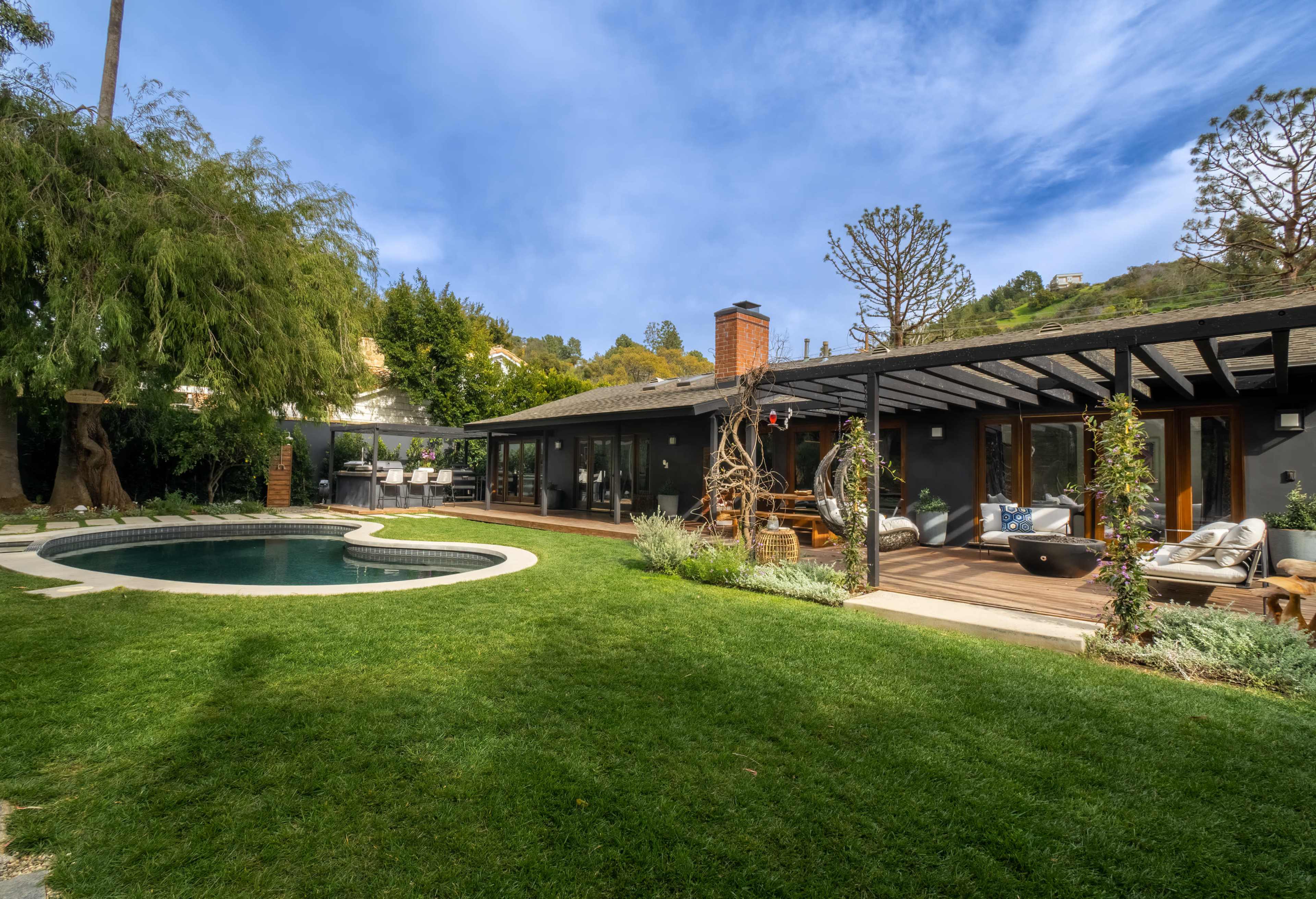 A modern house with a landscaped lawn and pool features large windows and a wooden deck, surrounded by trees and hills.