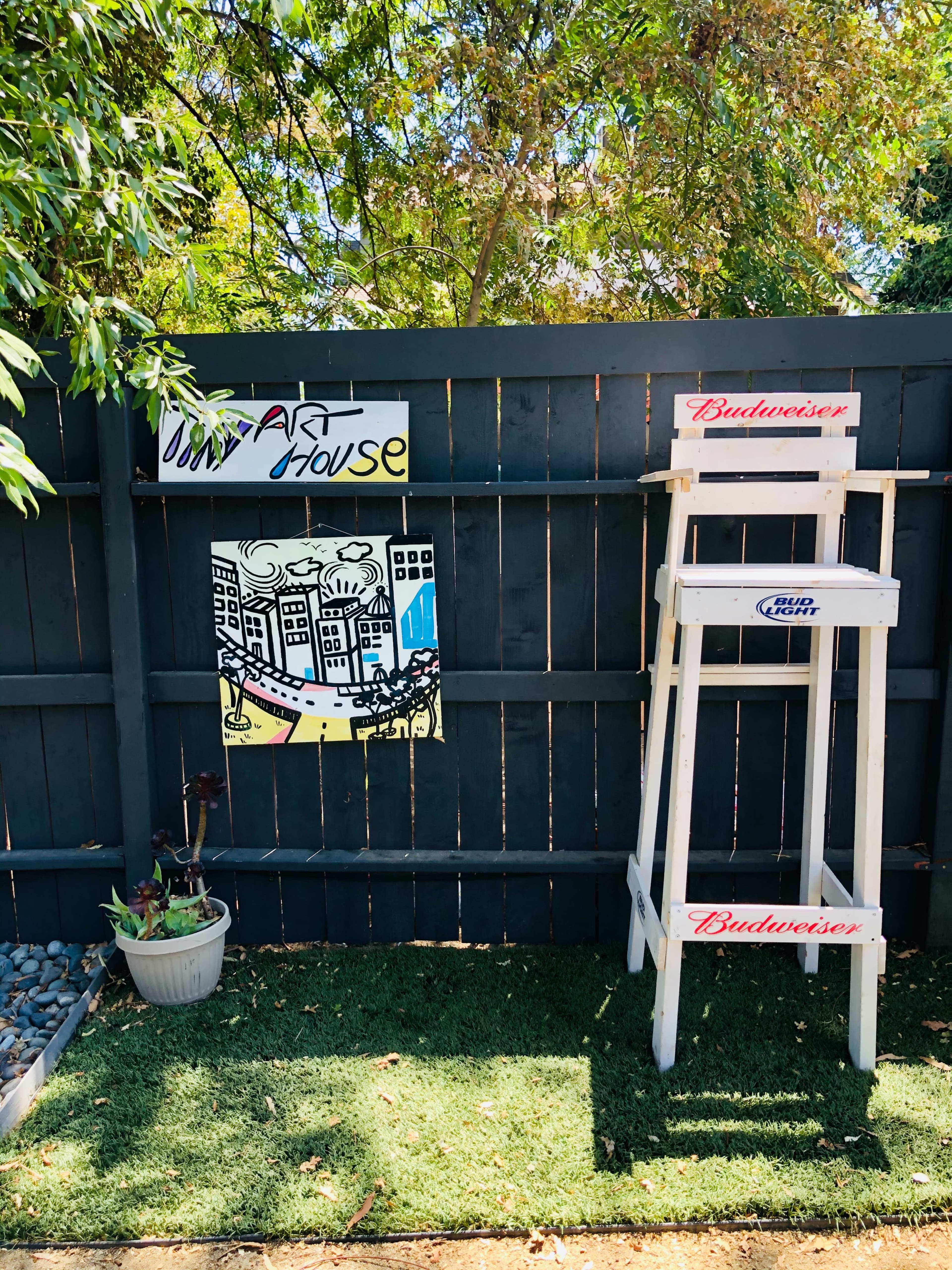 A wooden fence displays a colorful art piece and a tall white stool beside a small potted plant on a grassy area.