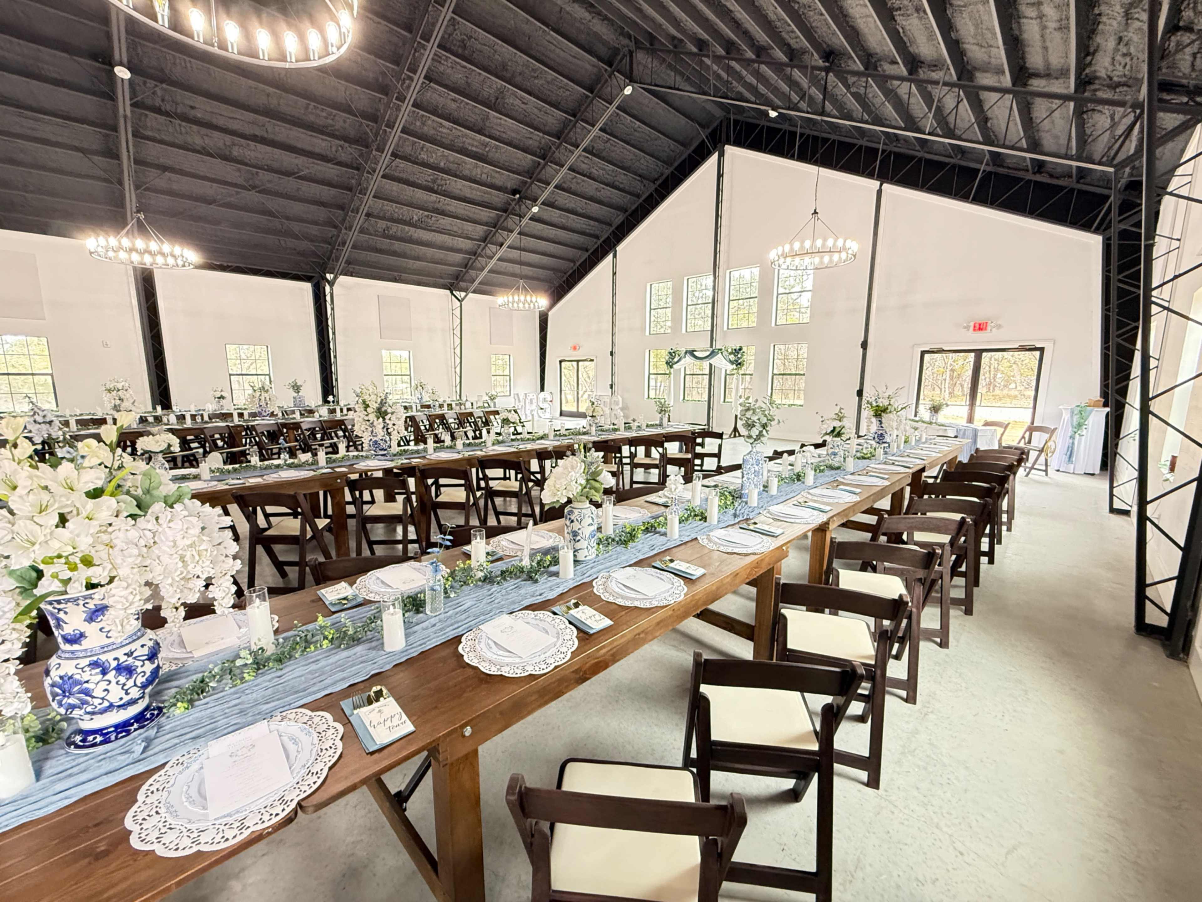 A spacious indoor banquet hall features long wooden tables set with white tableware and floral centerpieces, surrounded by elegant chandeliers.