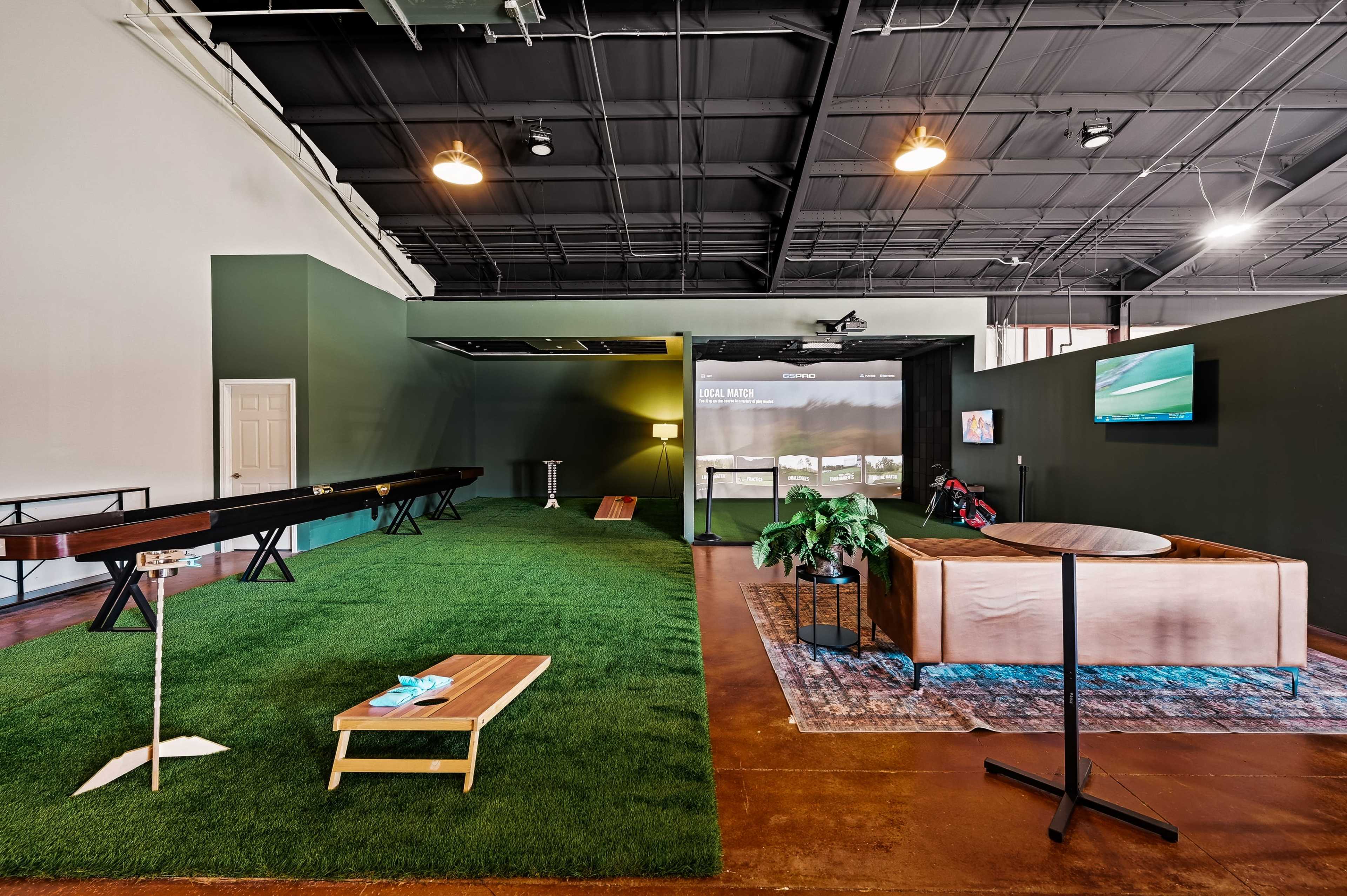 The image shows an indoor recreational space featuring a putting green, a lounge area with a sofa, a coffee table, and a television mounted on the wall.