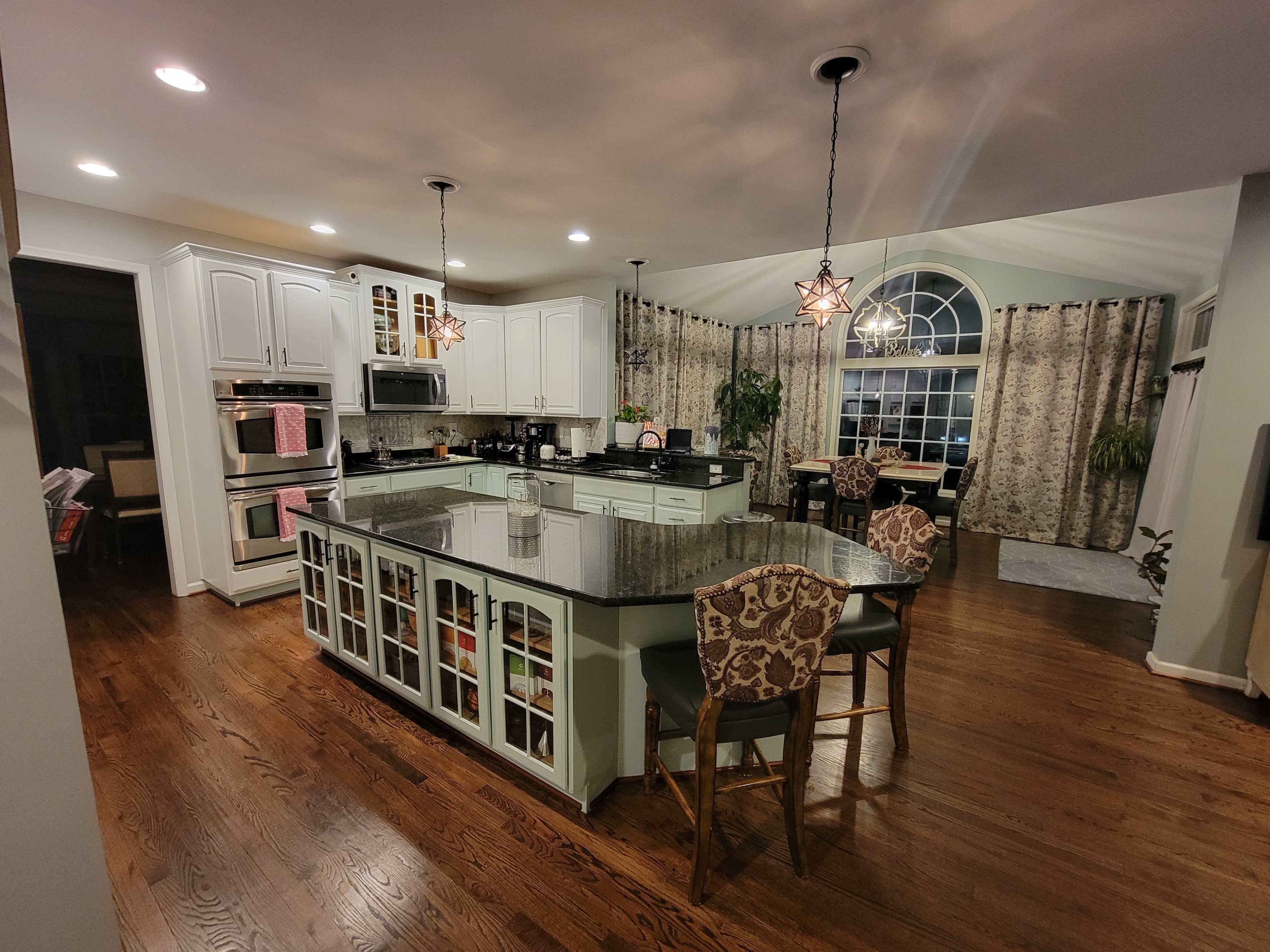 A spacious kitchen with a large island, dark wood flooring, white cabinetry, and a large window with ornate curtains.