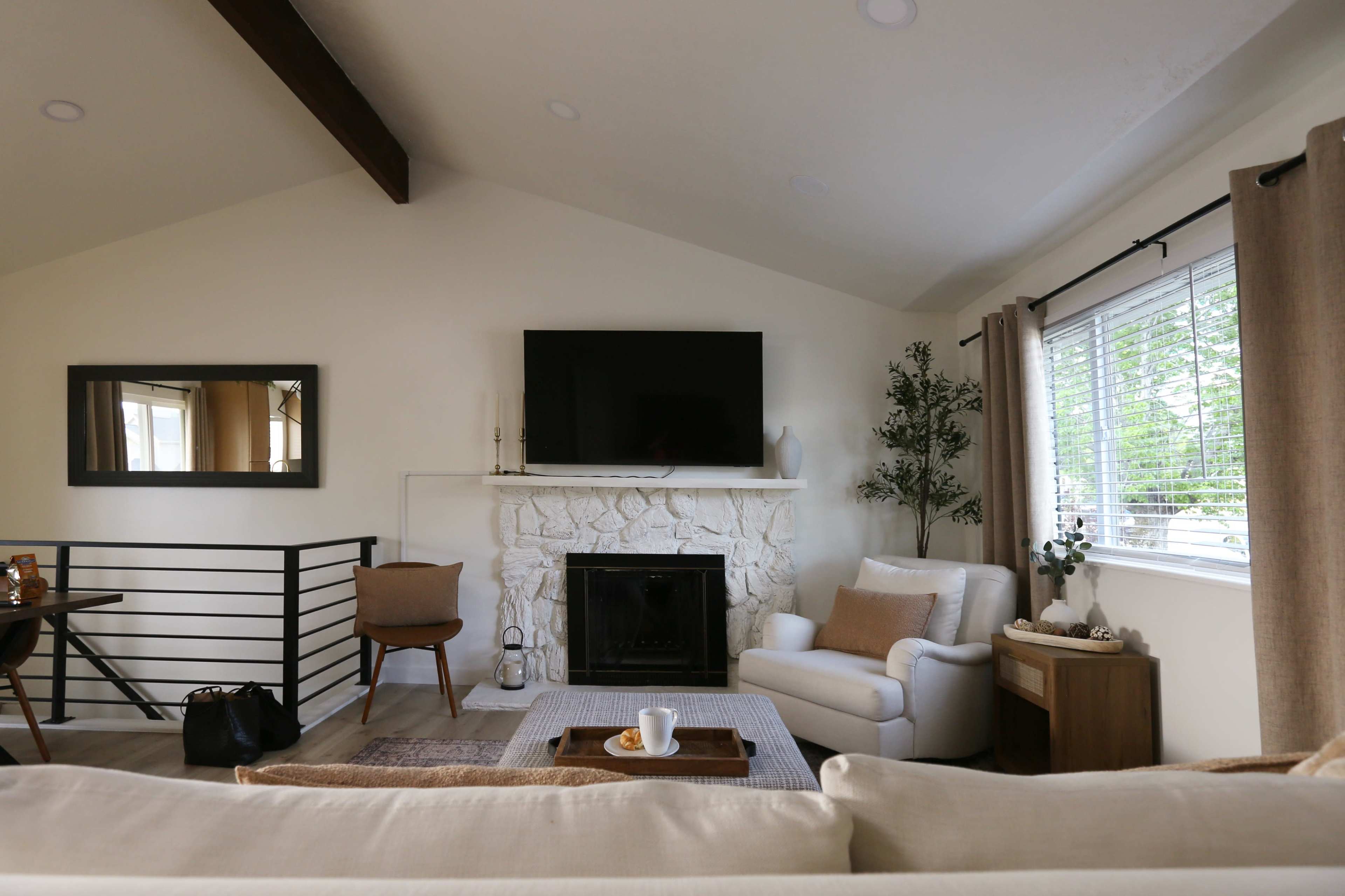 The image shows a cozy living room featuring a white stone fireplace, a mounted television, a comfortable armchair, and a small coffee table with a cup and snacks.