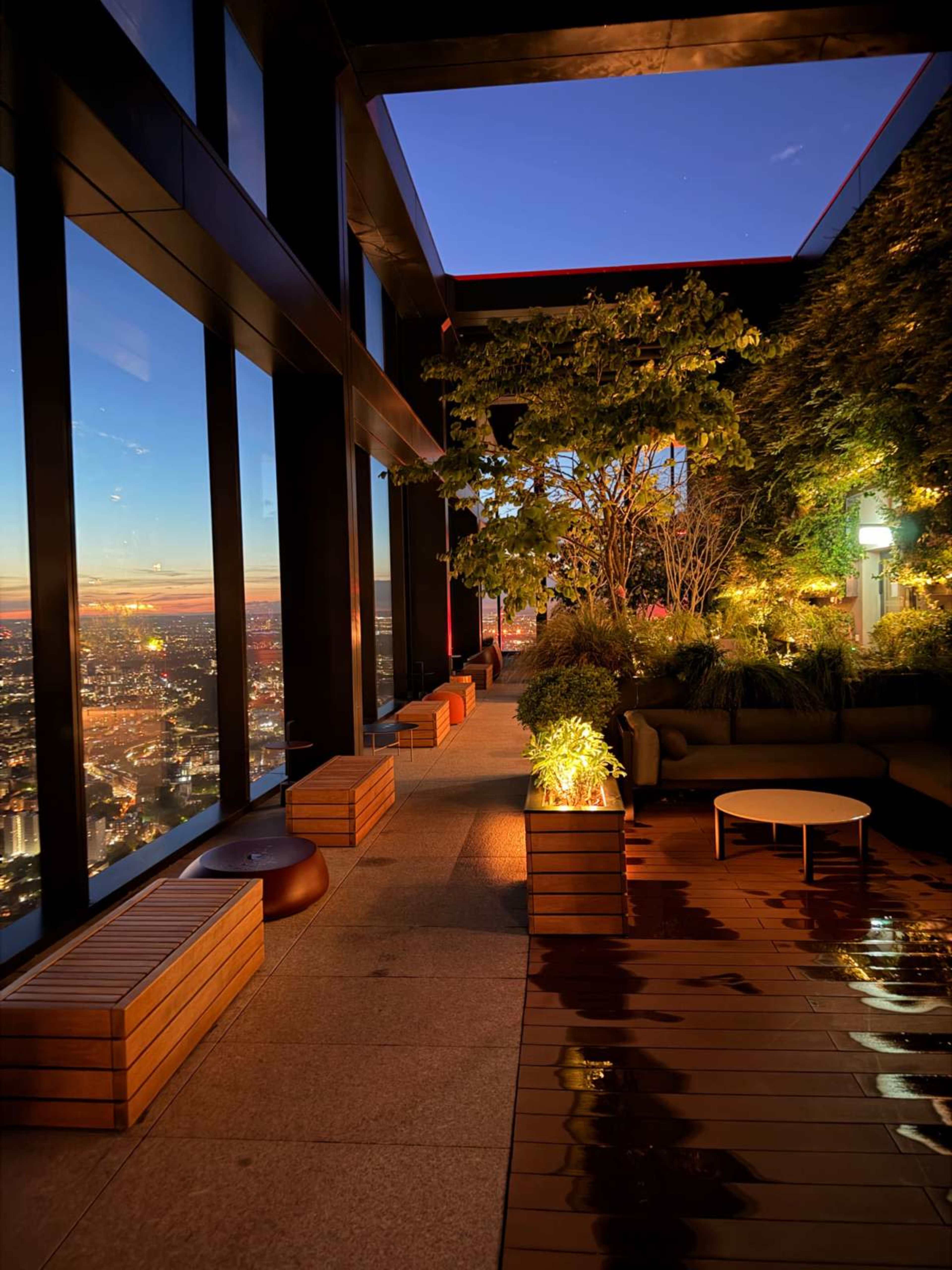 The image shows a rooftop terrace with wooden planters, seating areas, and a city skyline view at dusk.