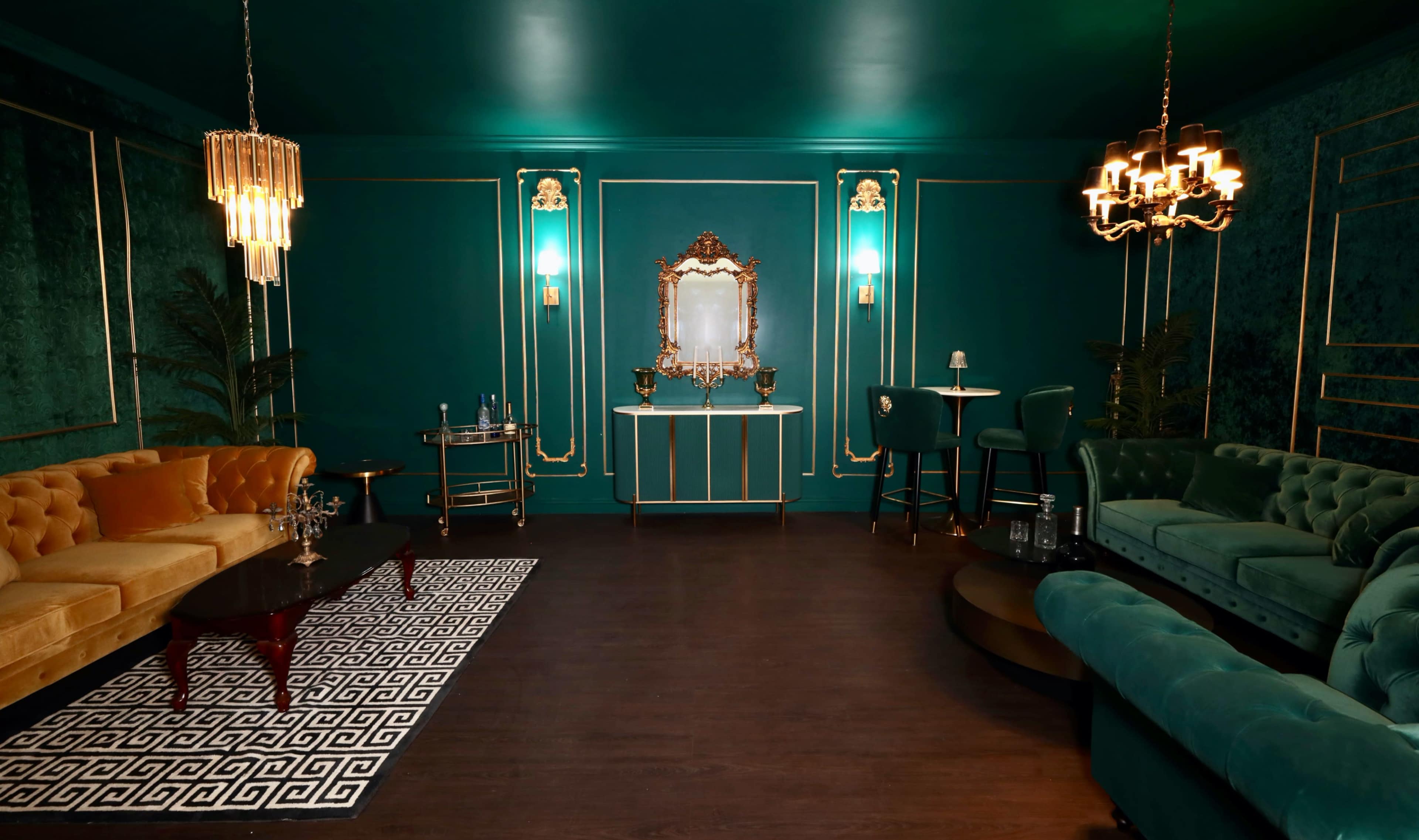 A luxurious lounge features teal walls, plush velvet furniture, elegant lighting fixtures, and a decorative mirror framed in gold.