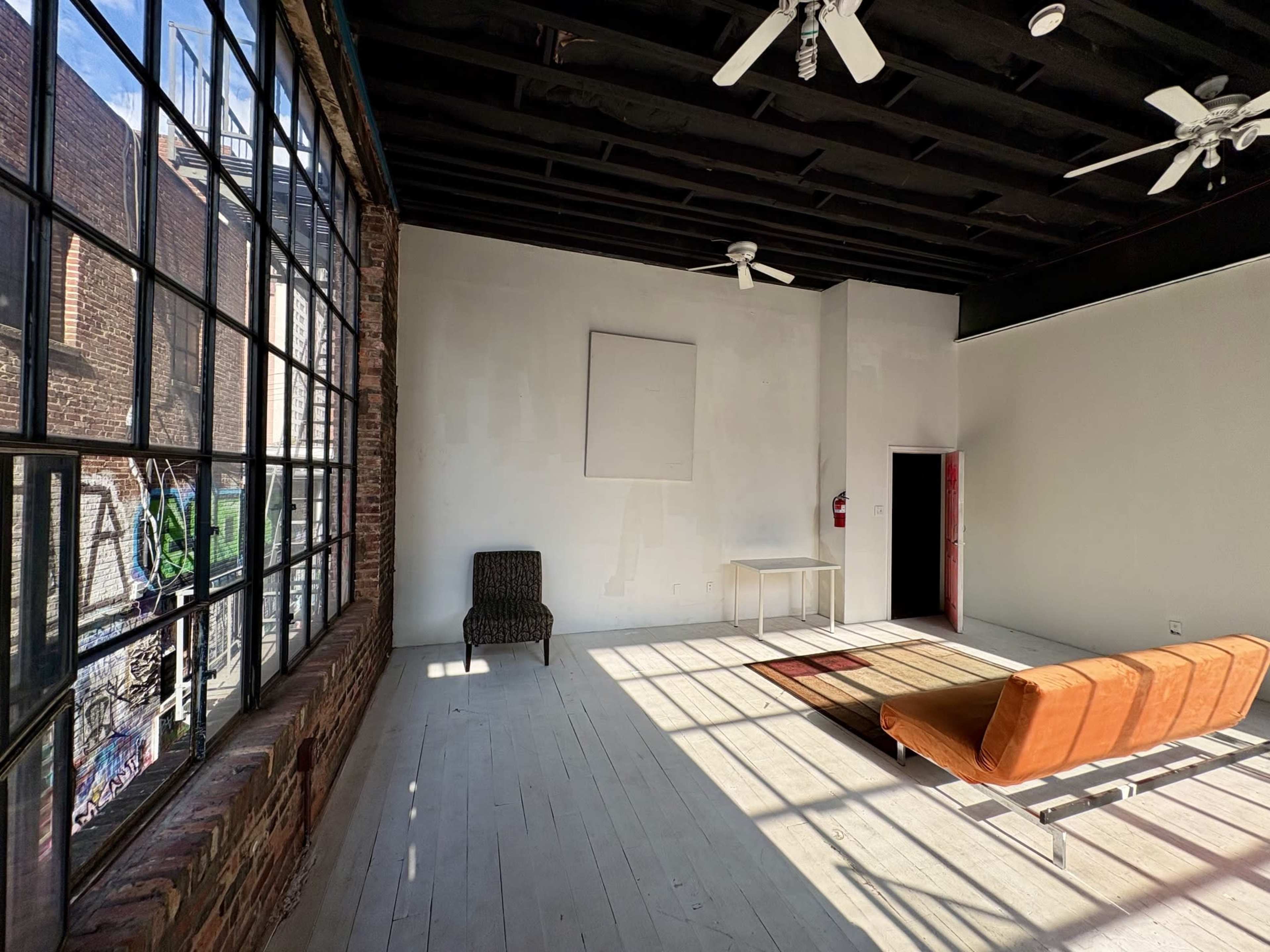 The image shows an empty, sunlit room with large windows, a brown chair, a table, and an orange couch on a wooden floor.