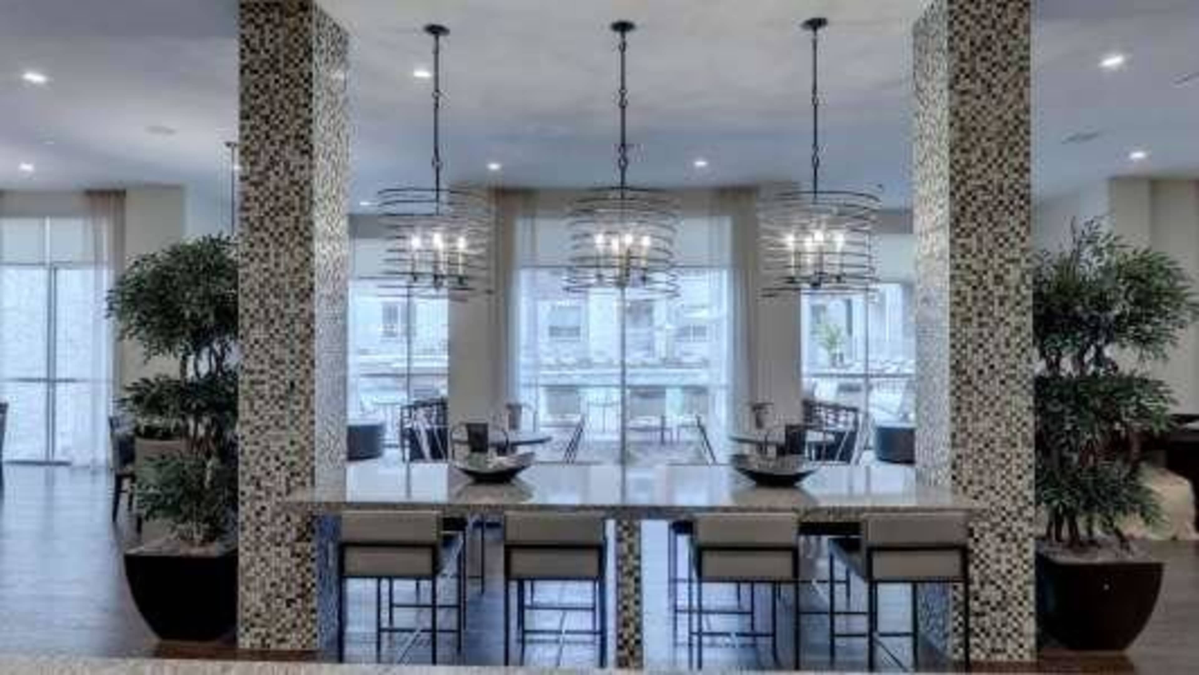 The image shows a modern dining area with a long table surrounded by chairs, elegant light fixtures overhead, and large windows allowing natural light to illuminate the space.