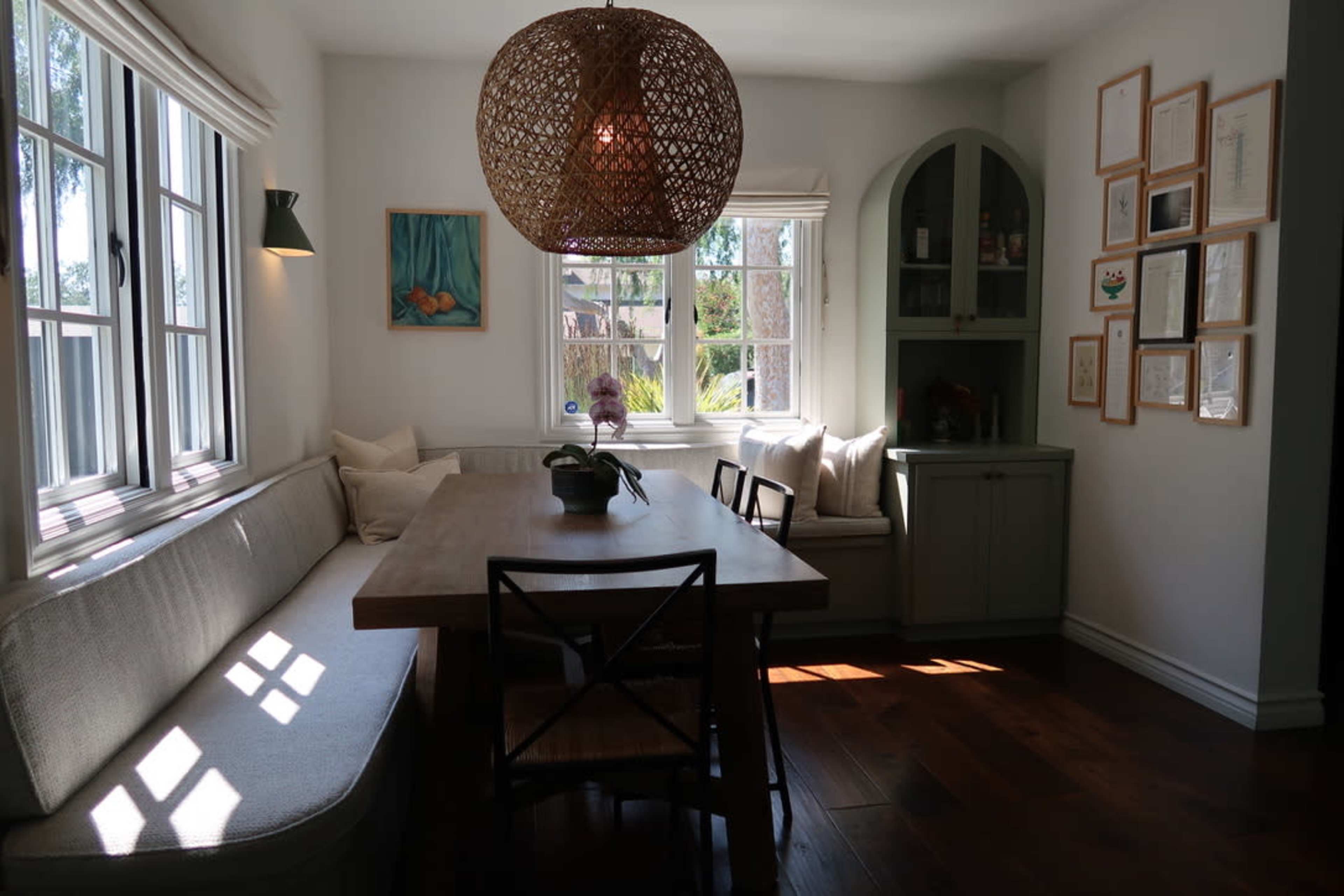 A dining area features a wooden table surrounded by chairs, with a cozy bench, large windows, and a woven pendant light.