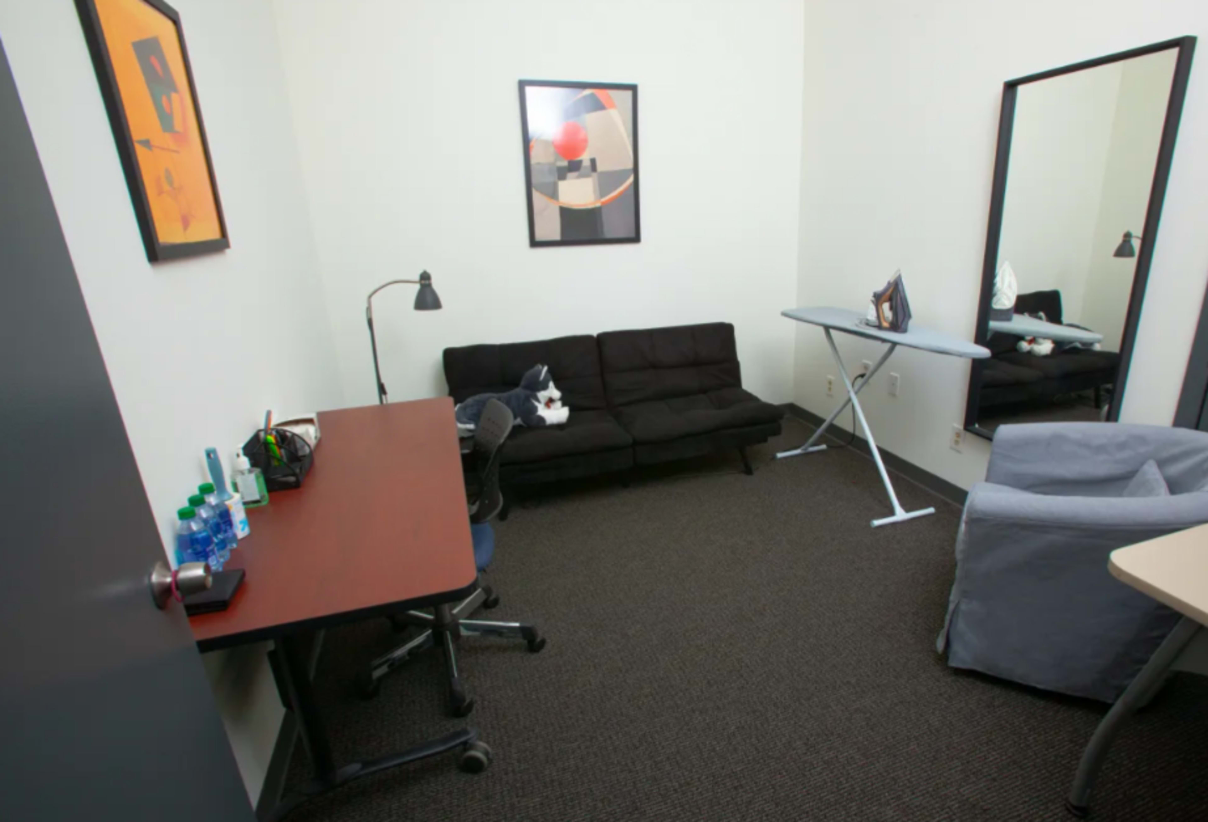 A small room with a black sofa, a desk with bottles, an ironing board, a mirror, and a blue armchair.