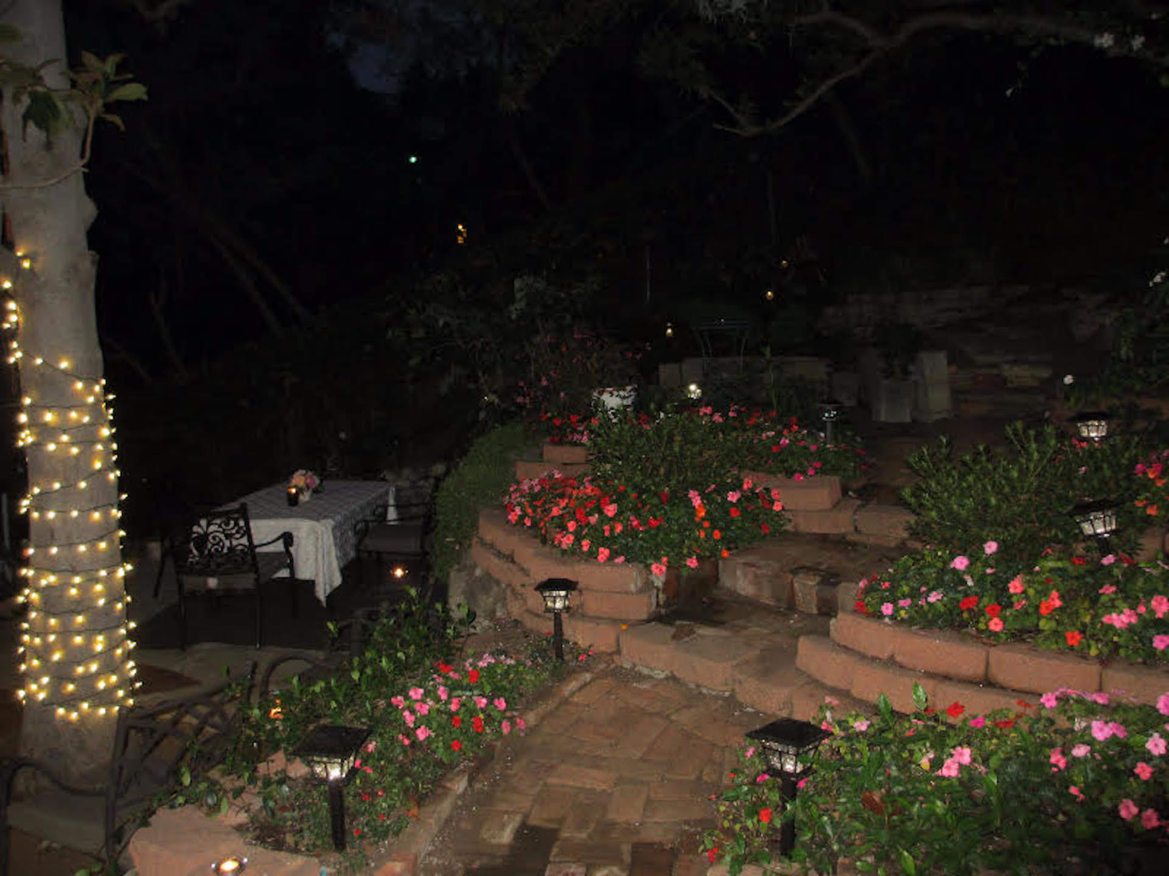 A dimly lit garden pathway with brick steps surrounded by flower beds and decorative lights leading to a small dining area.