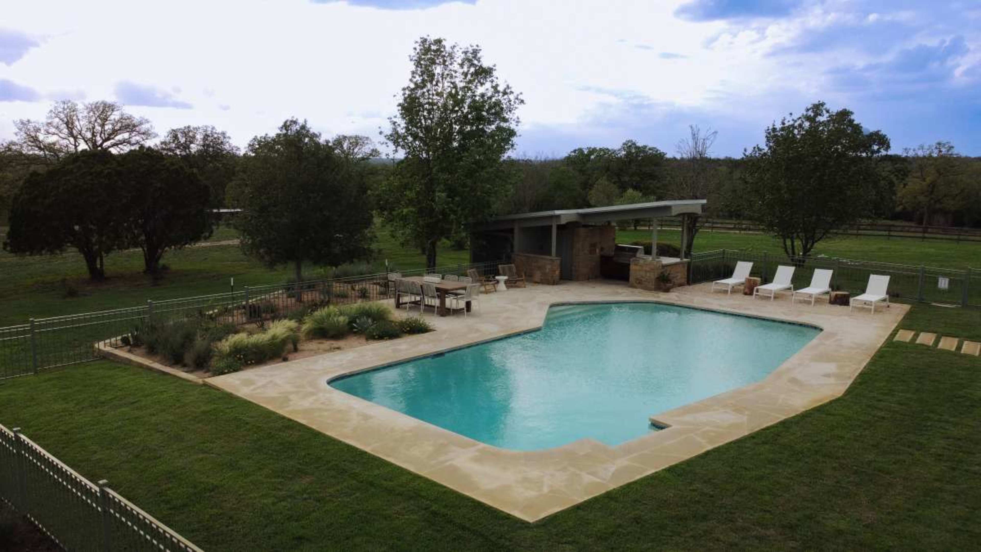 The image shows a rectangular swimming pool surrounded by a patio and lounge chairs, set in a grassy area with trees in the background.