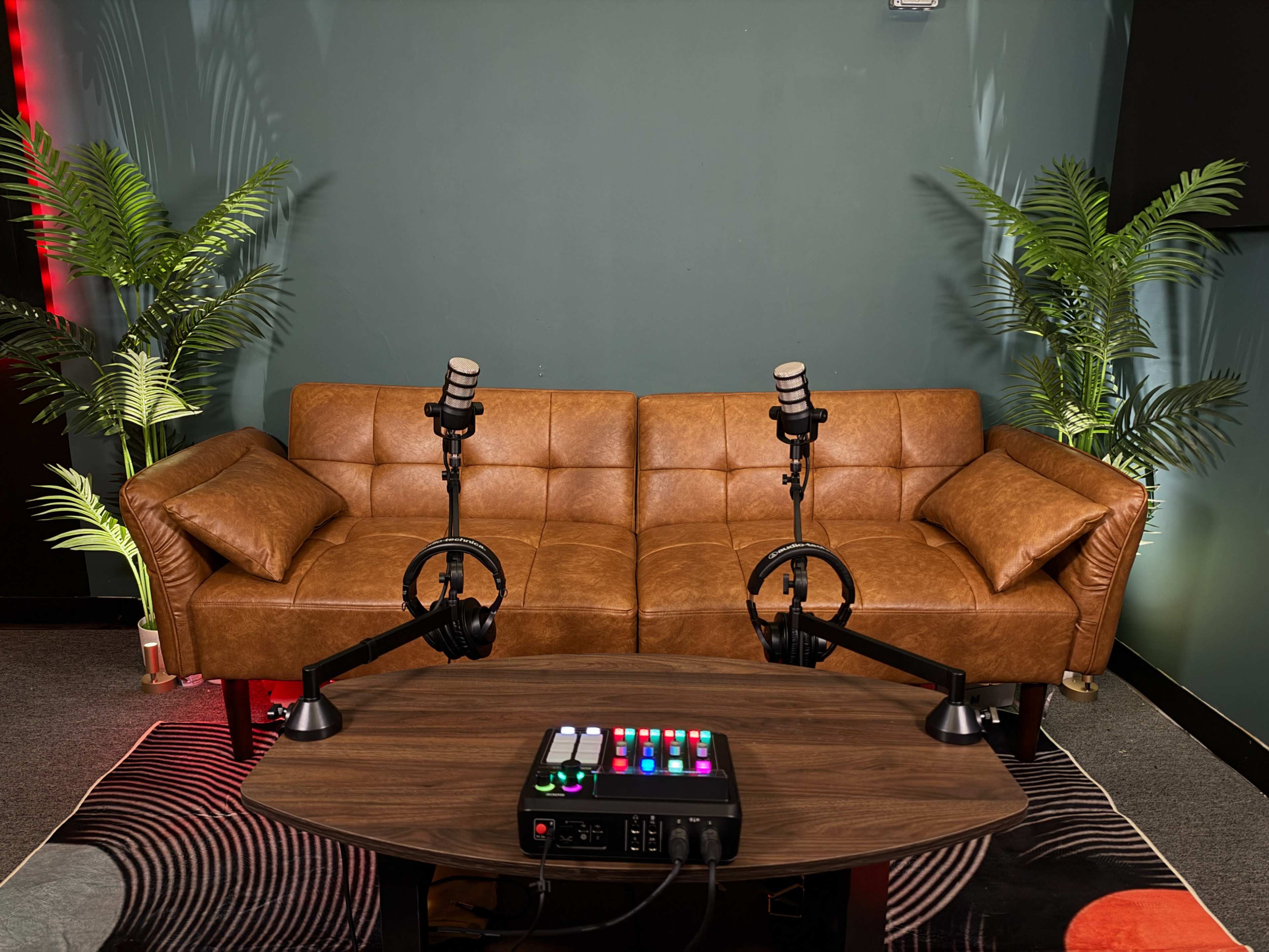 The image features a cozy brown leather sofa flanked by two microphones and a sound mixing device on a coffee table, set against a green wall with decorative plants.