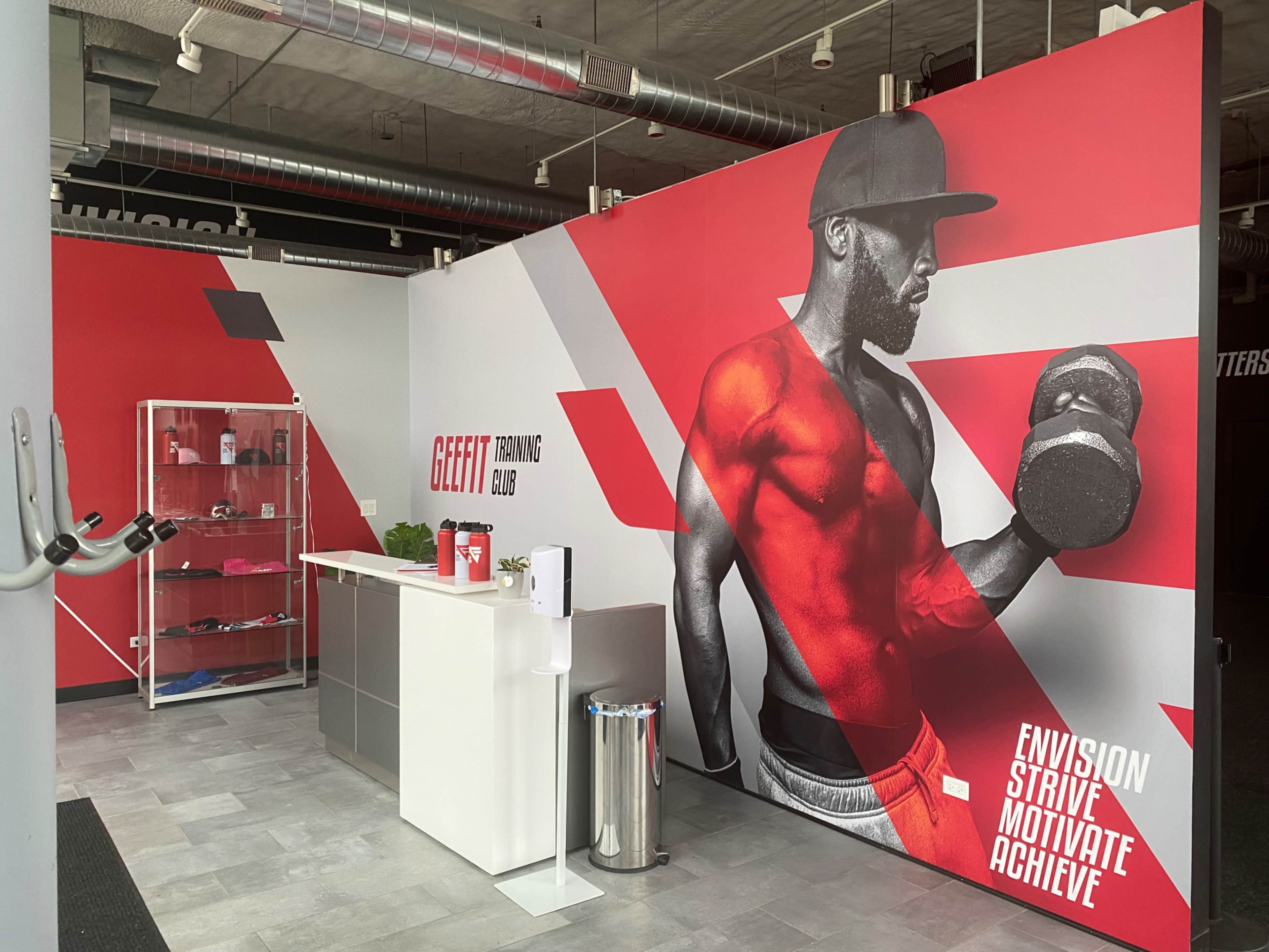 A modern gym reception area featuring a large wall mural of a muscular man holding a dumbbell and a sleek front desk with promotional materials nearby.