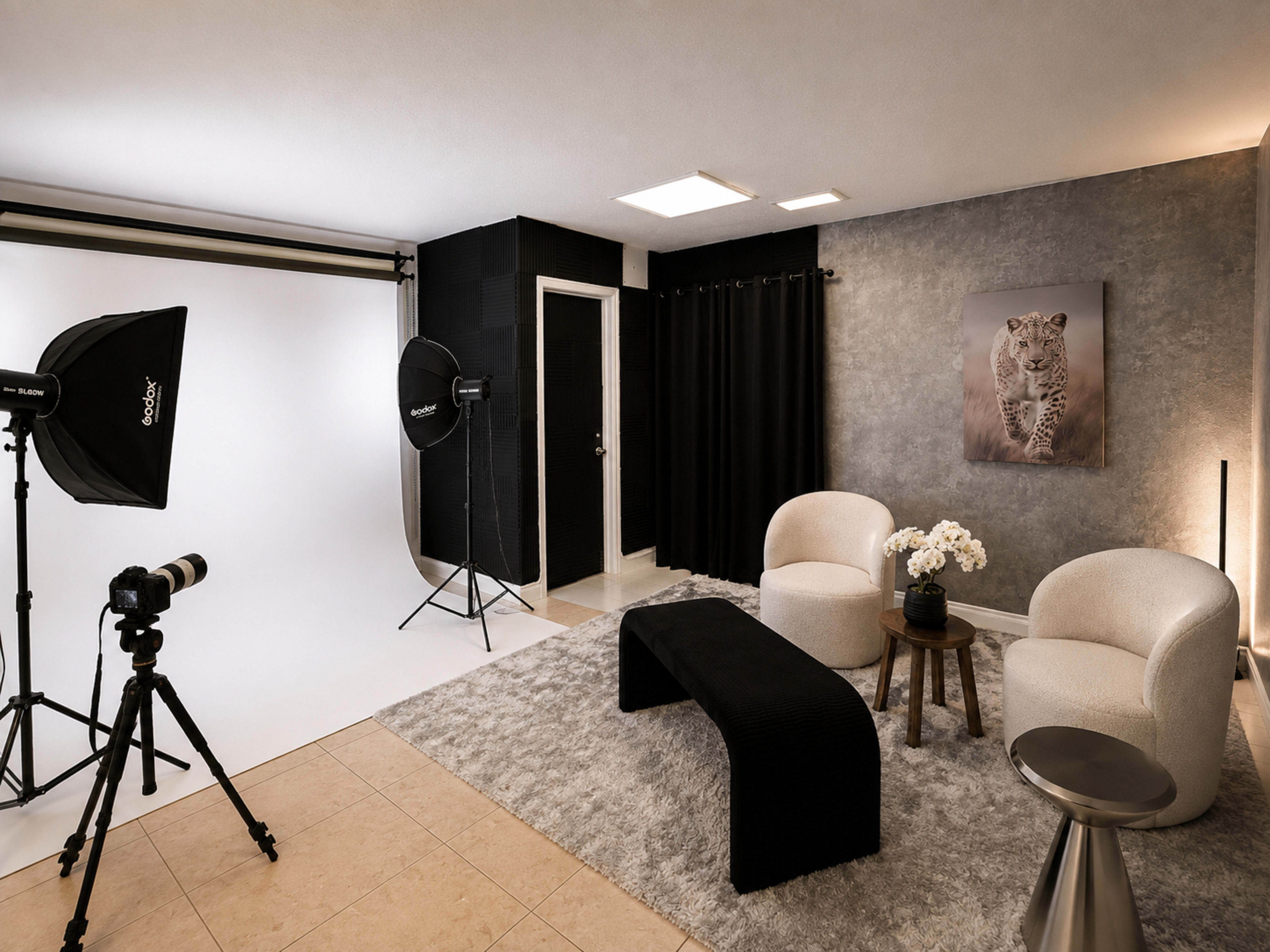The image shows a photography studio with a white backdrop, two upholstered chairs, a black coffee table, and lighting equipment.