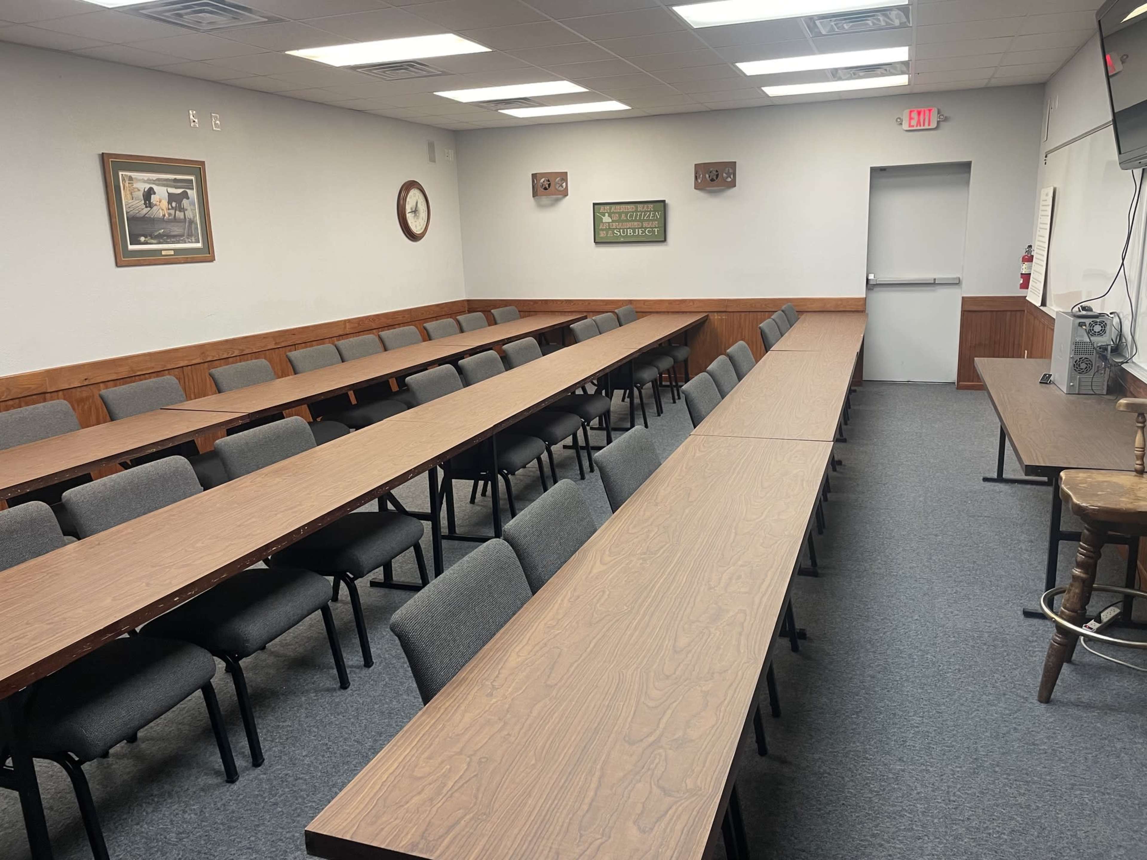 A spacious meeting room features several long tables arranged in rows, with grey chairs placed along each table and wall decorations including framed pictures and signs.
