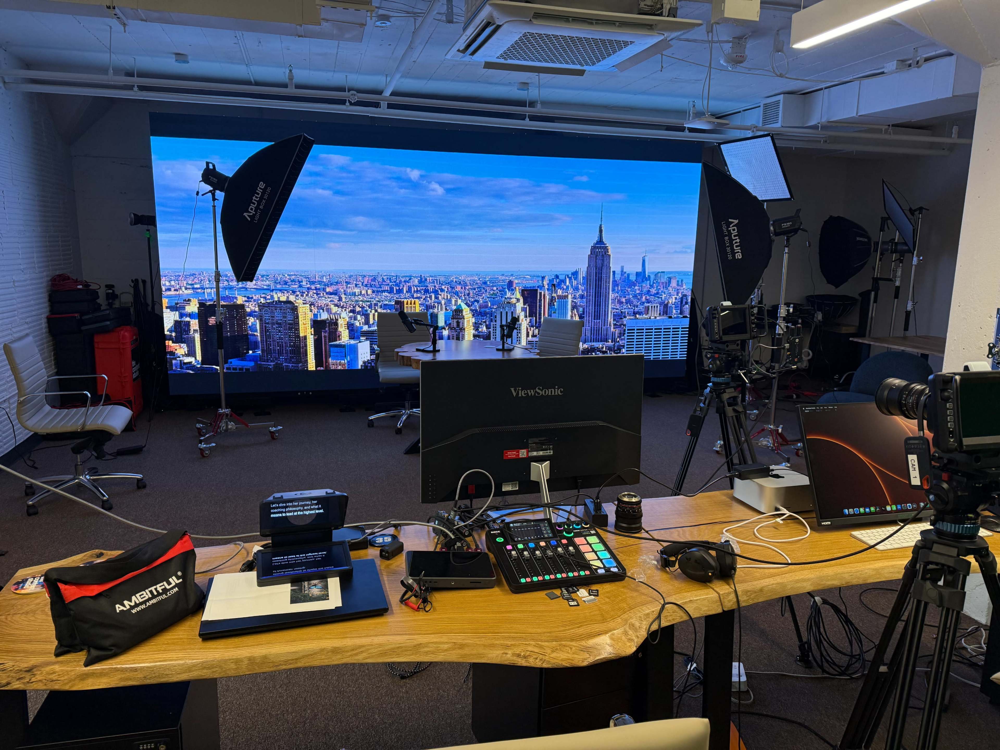 A modern studio setup features a large backdrop of a city skyline, equipped with cameras, lighting equipment, and a central workstation with multiple monitors and control panels.