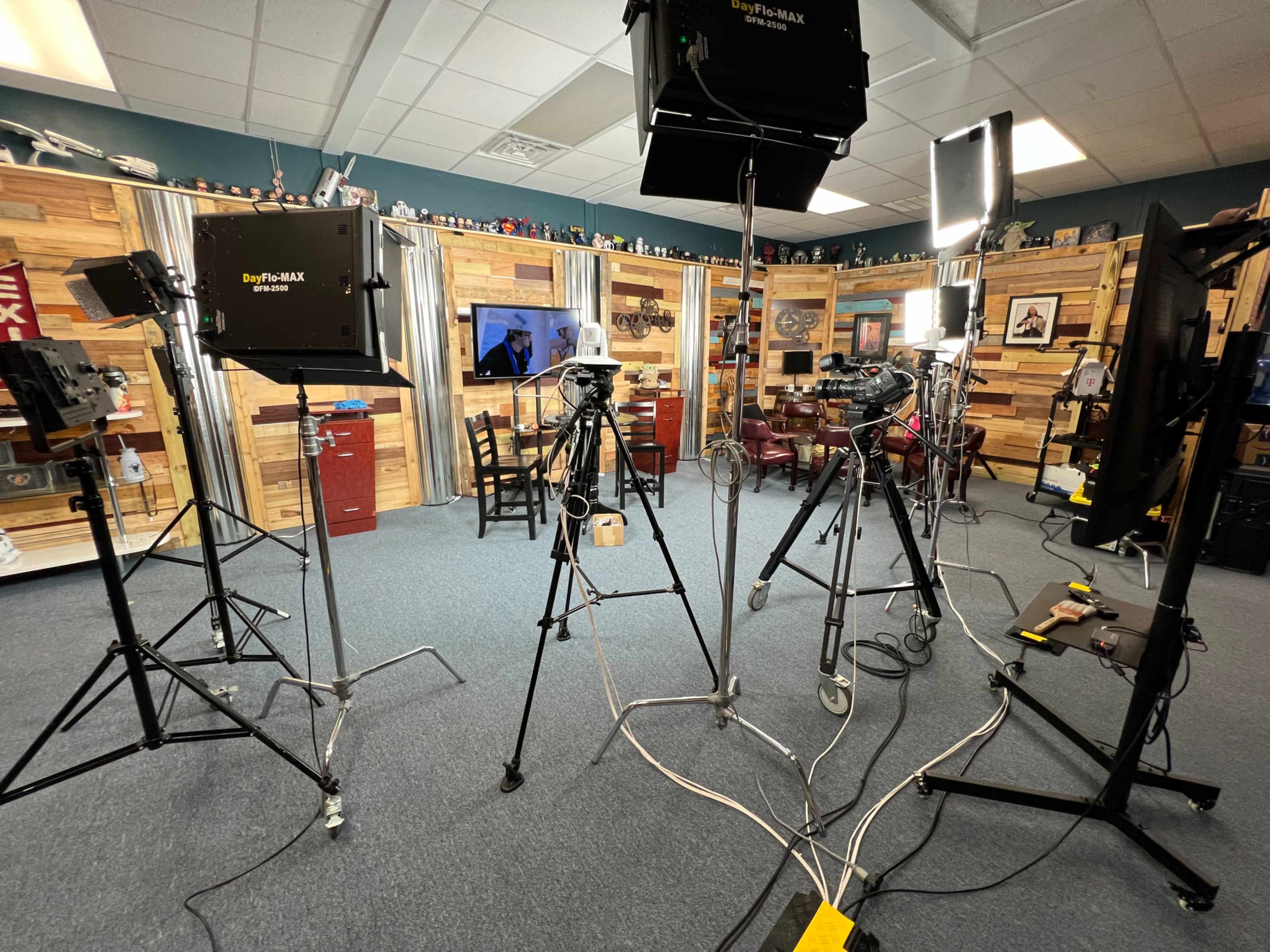 The image shows a studio setup with several cameras on tripods, bright lights, and a television screen in a room decorated with wooden panels.