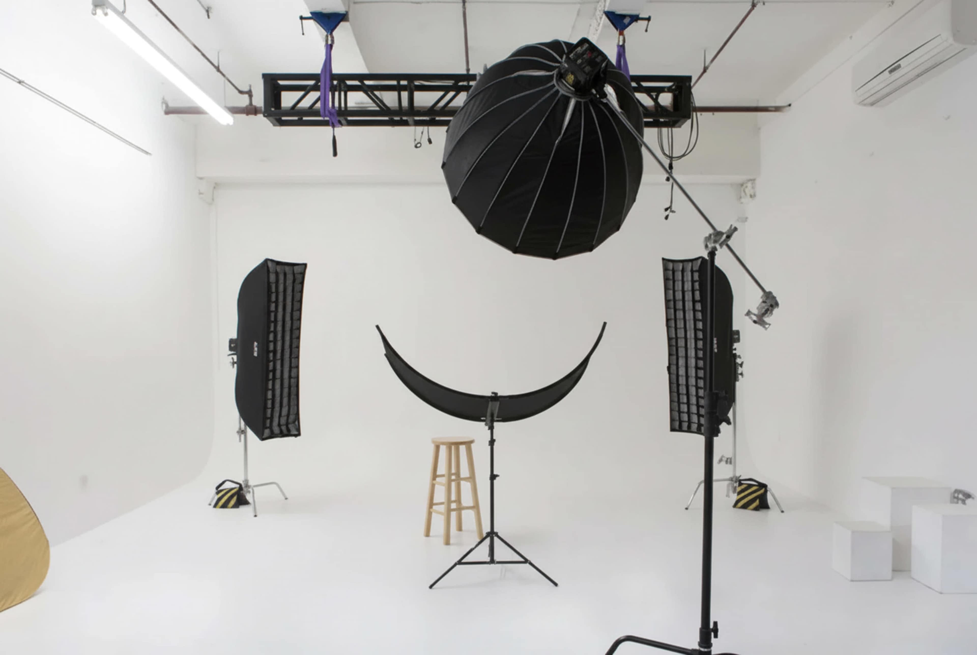 The image shows a photography studio featuring a central stool, lighting equipment, and large softboxes set against a white backdrop.