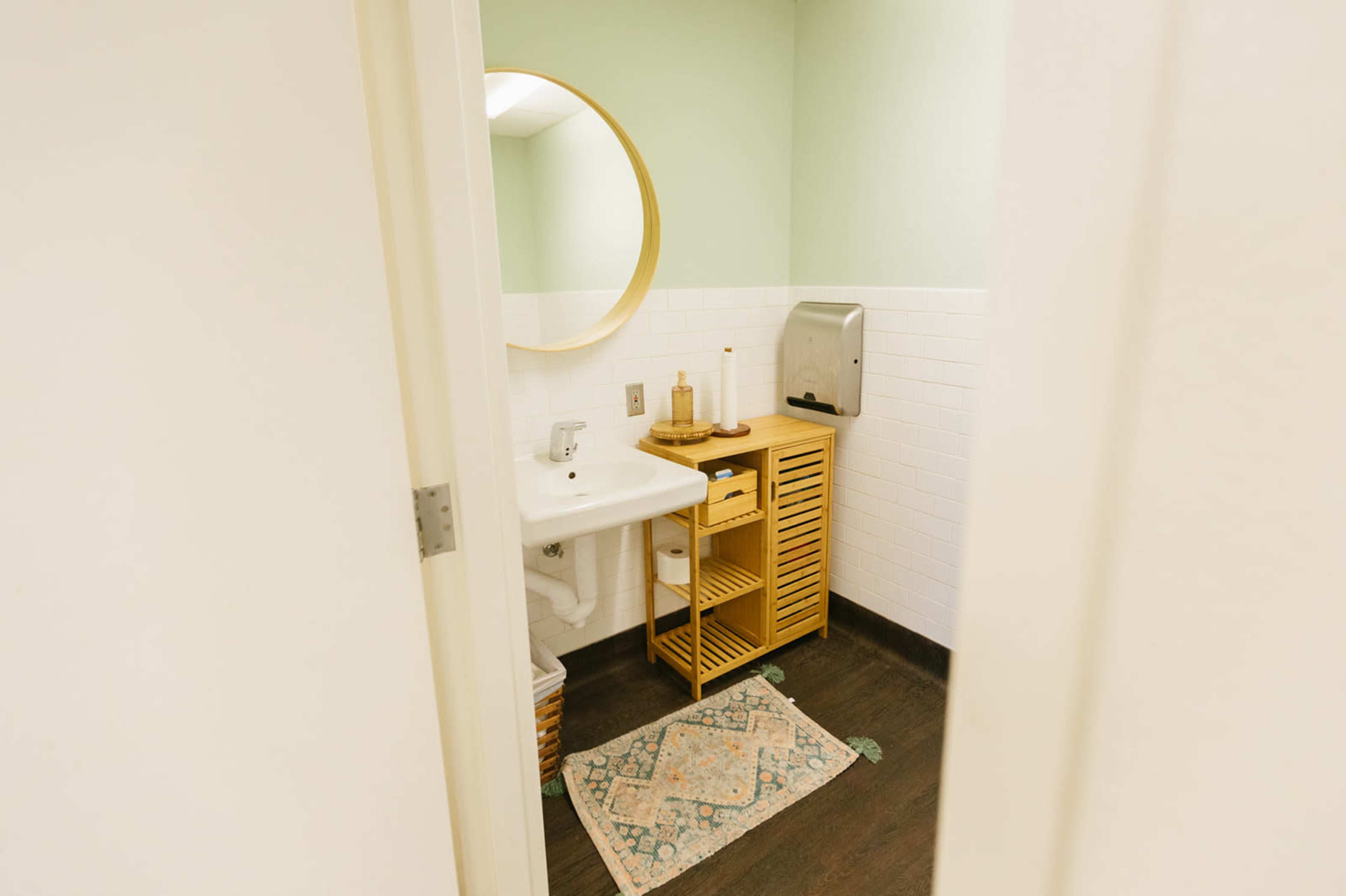 A small bathroom with a sink, a round mirror, a brown wooden storage unit, and a hand dryer on a green and white tiled wall.