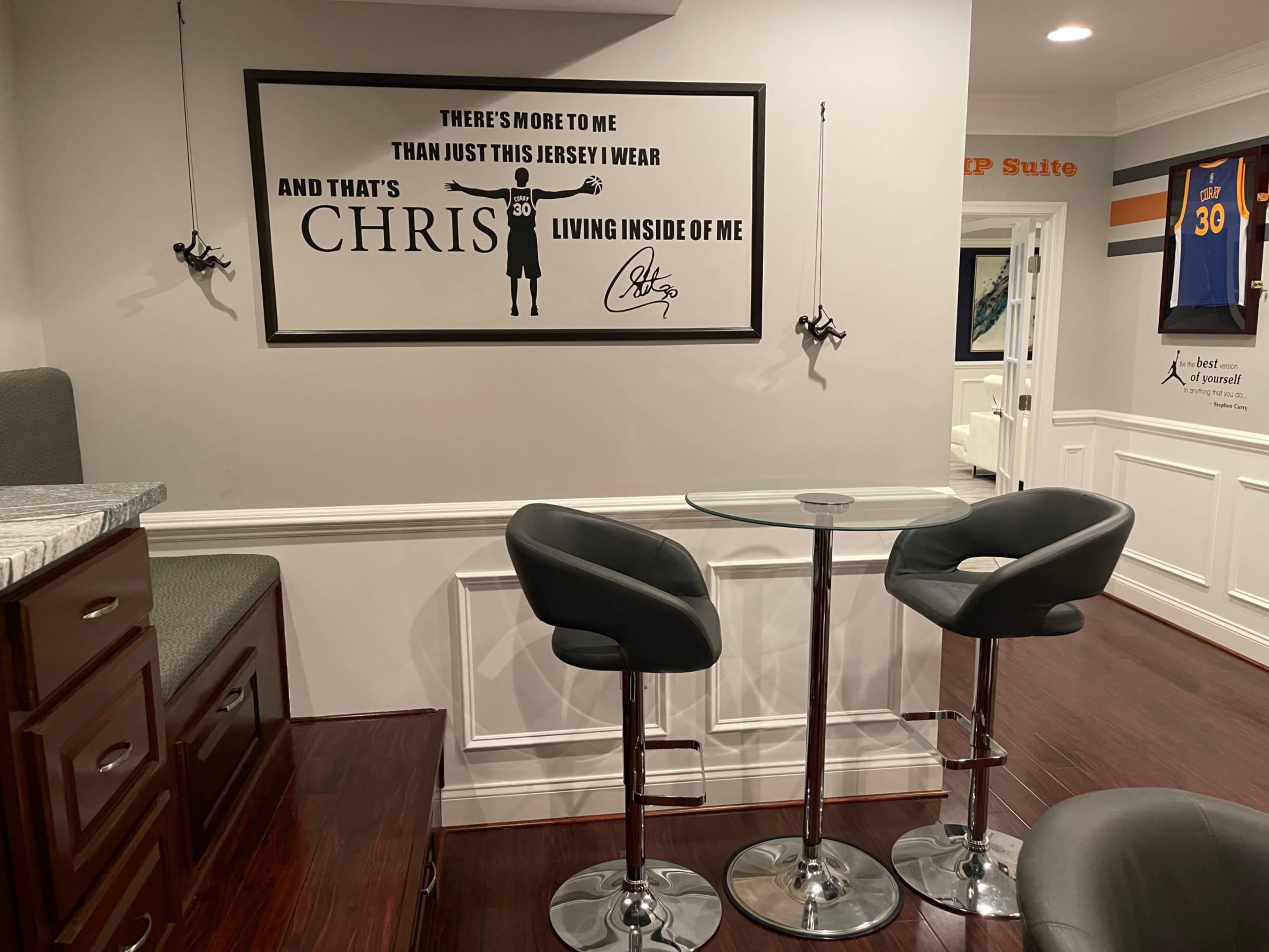 A small seating area with a glass table and two black bar stools, surrounded by white walls featuring a framed wall art depicting a jersey and the name "CHRIS."