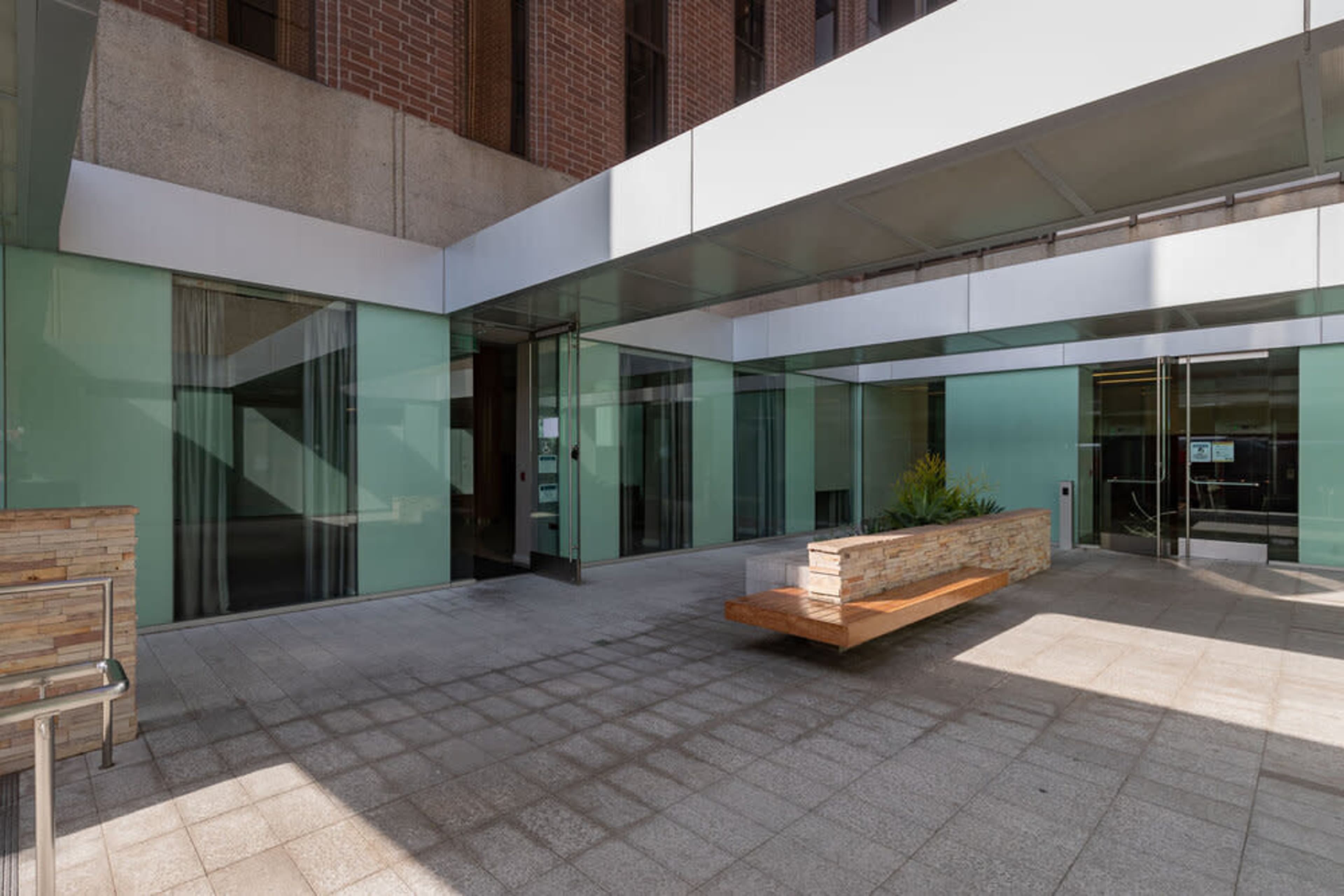 The image shows a modern entrance area featuring large glass walls, a wooden bench, and a landscaped planter in an open space.
