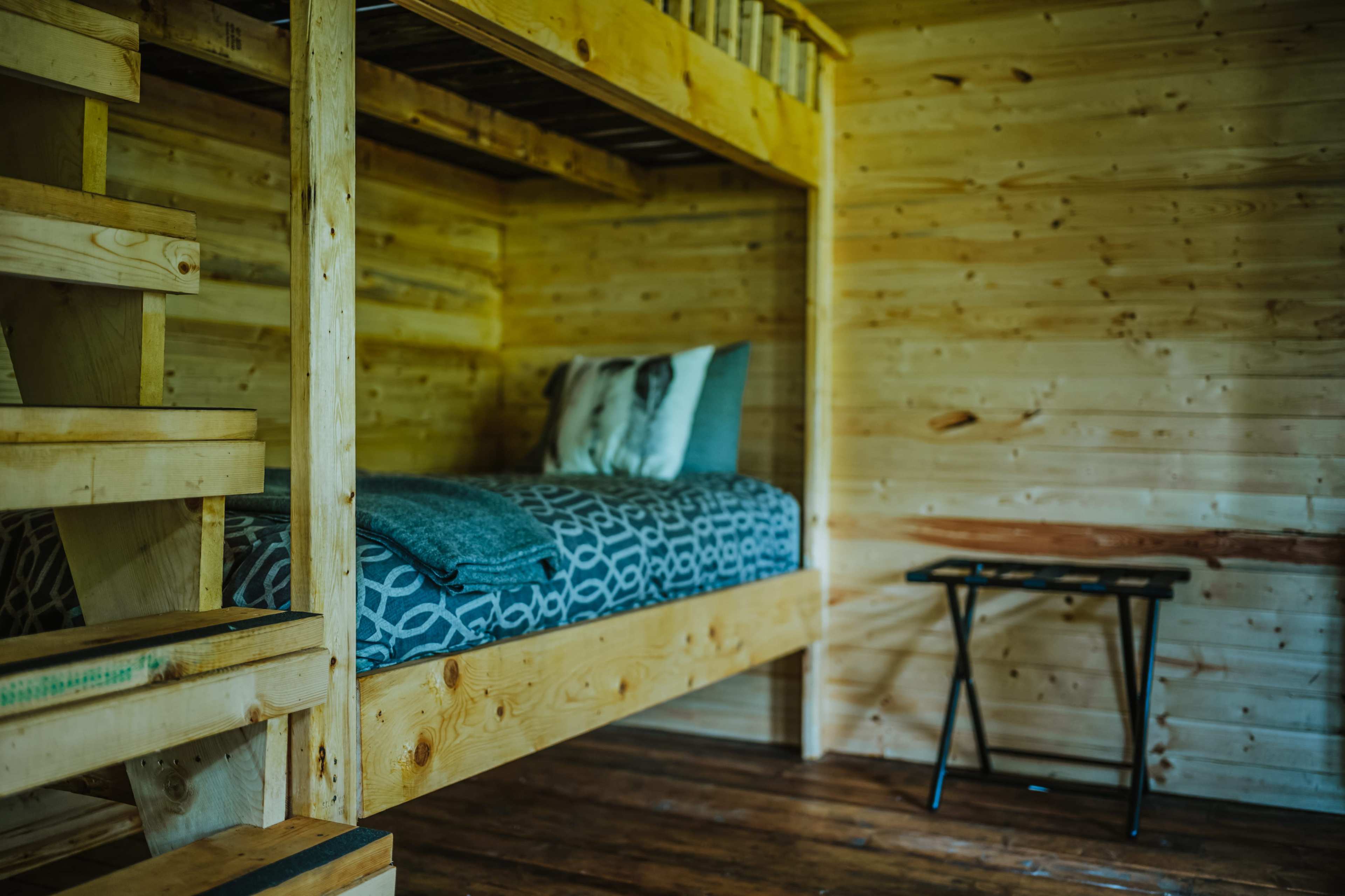 The image shows a wooden bunk bed with a single mattress and a small side table in a cabin-like room with wooden walls and floor.