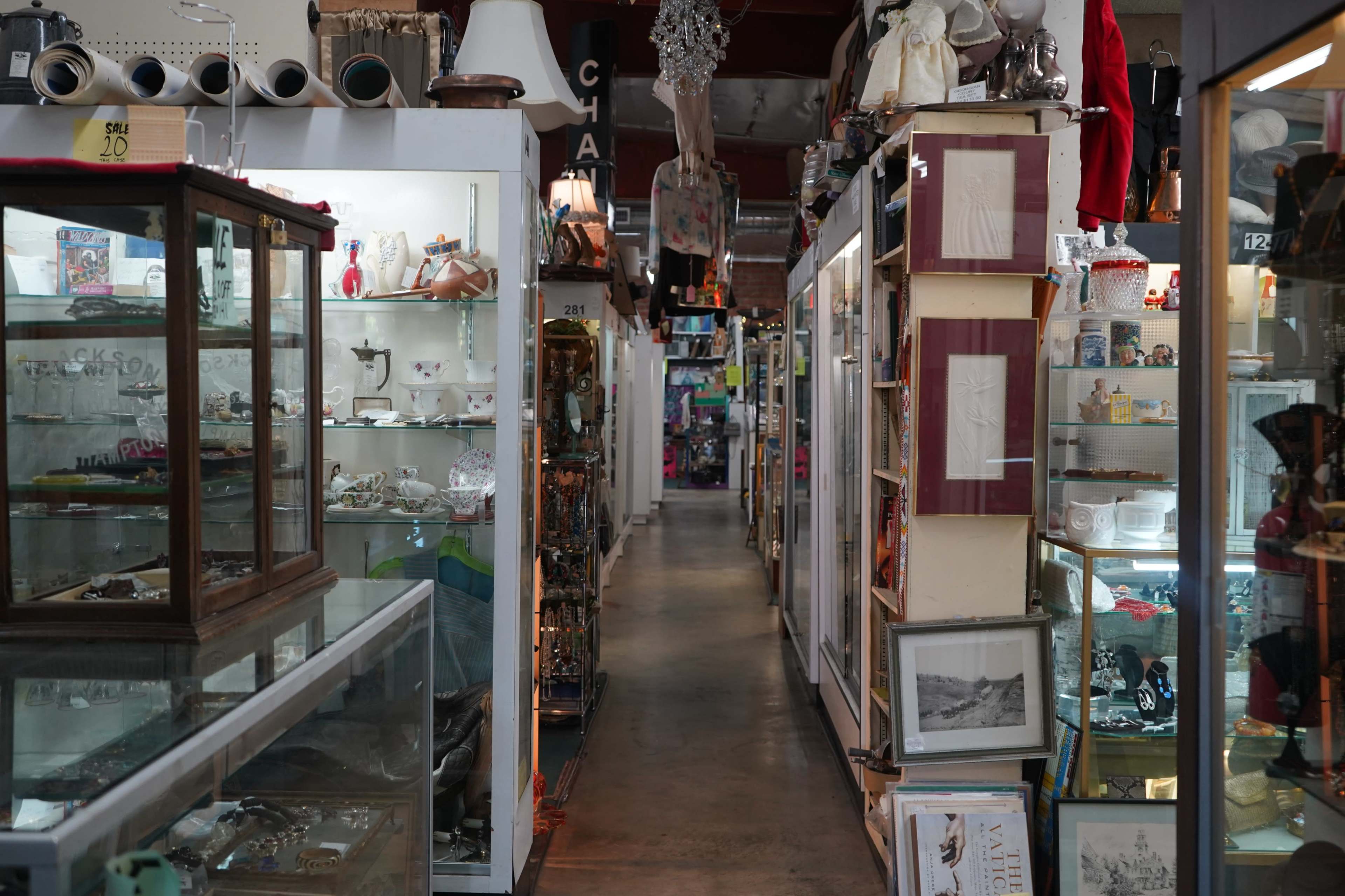 The image shows a narrow aisle in an antique shop lined with display cases filled with various collectibles and items for sale.