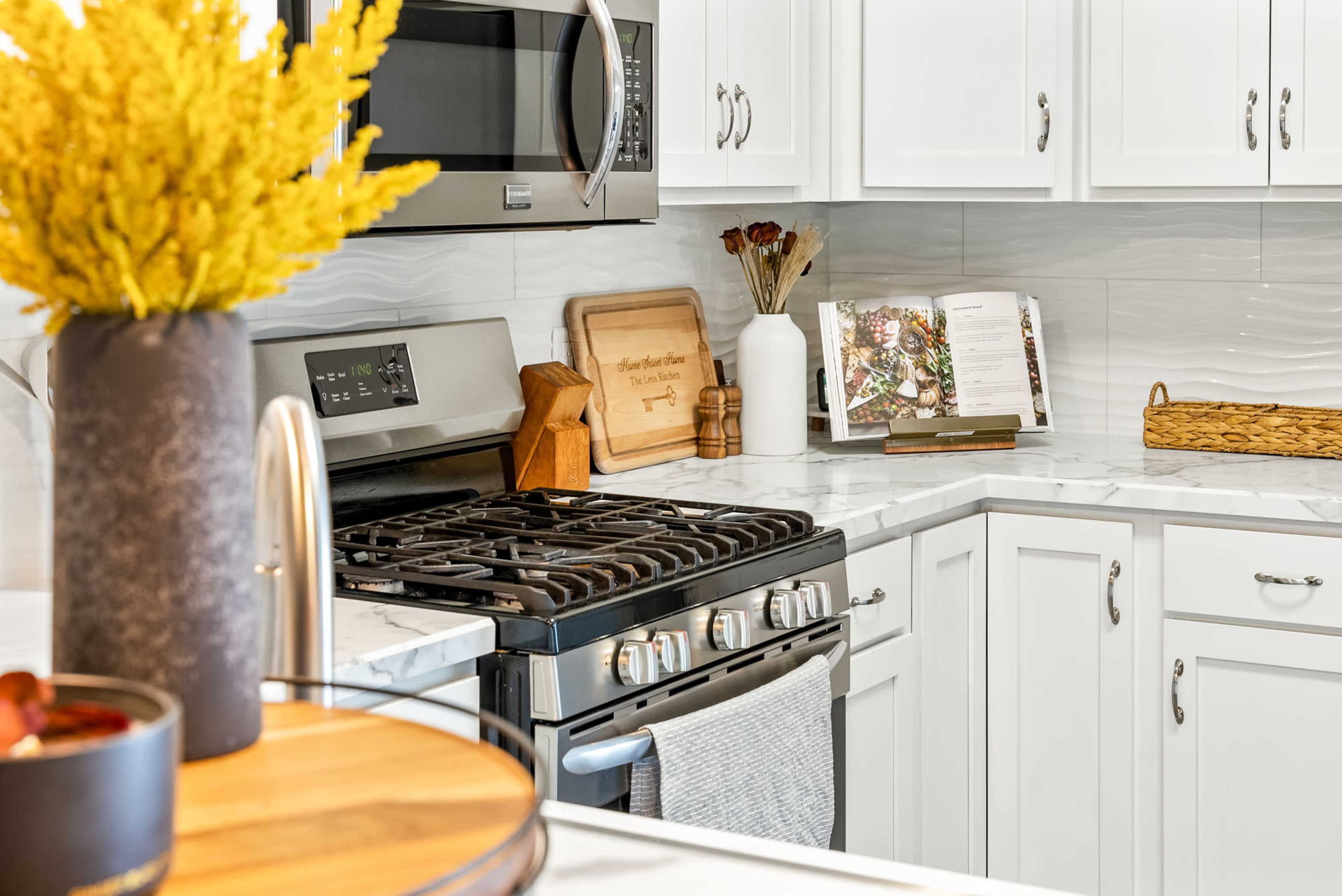 A modern kitchen features a stainless steel microwave, a gas stove with a light gray dish towel, and white cabinetry, with decorative items including a flower arrangement and a recipe book on the countertop.