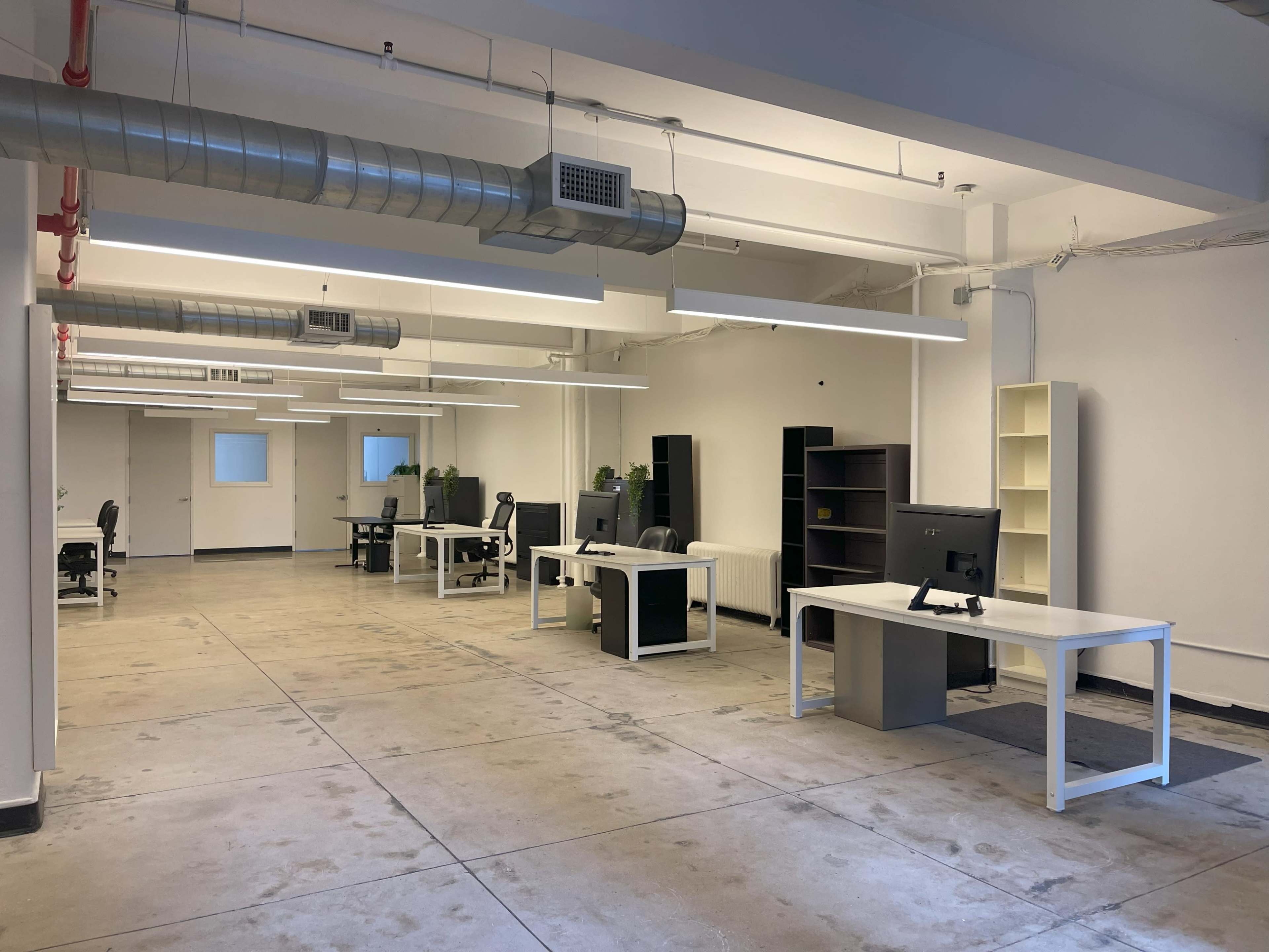 The image shows a spacious, minimally furnished office area with several desks, chairs, and computers arranged in an open layout.