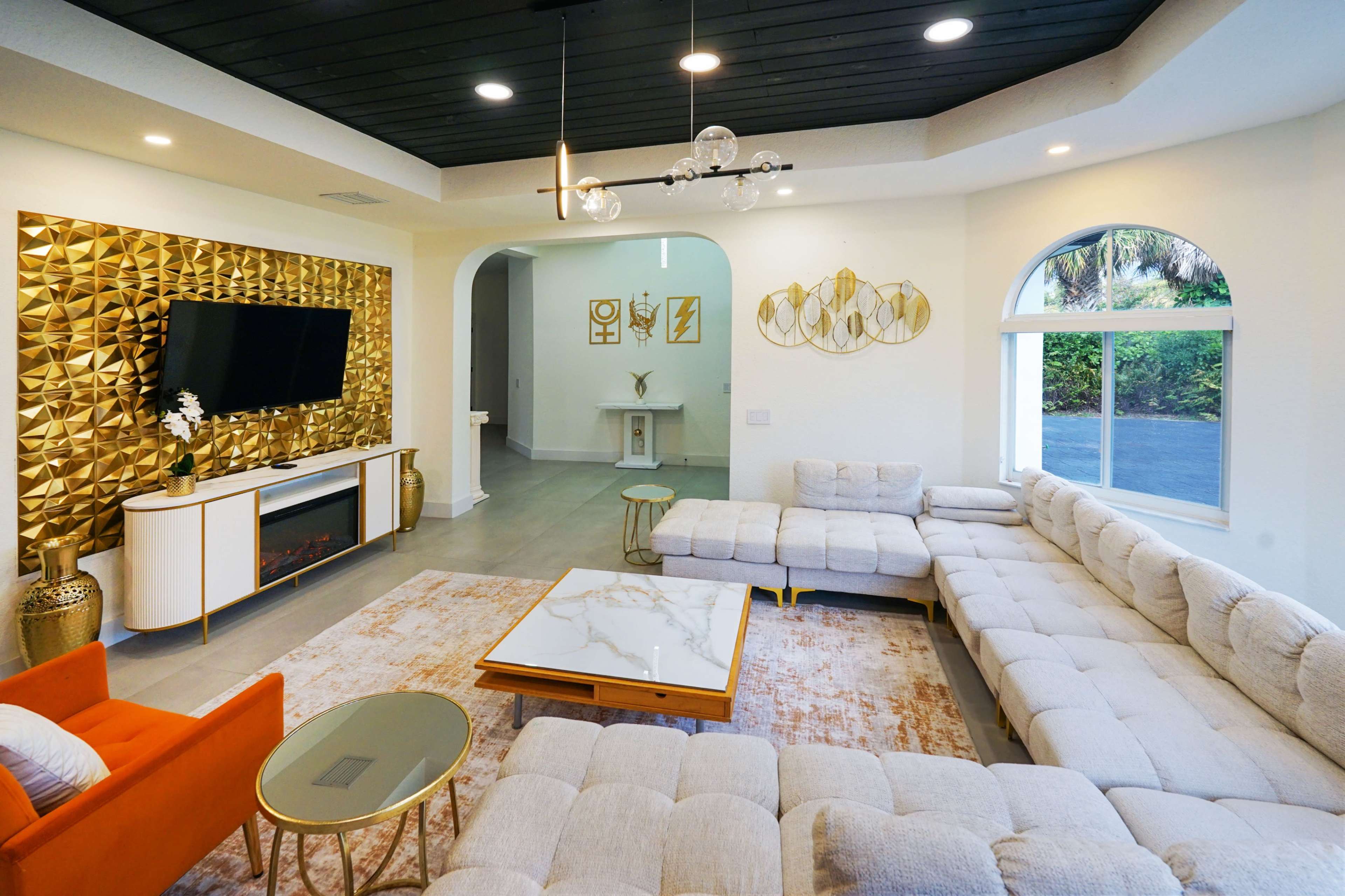 The image depicts a spacious living room featuring a sectional sofa, a coffee table, a large television mounted on a gold accent wall, and decorative elements in a modern style.