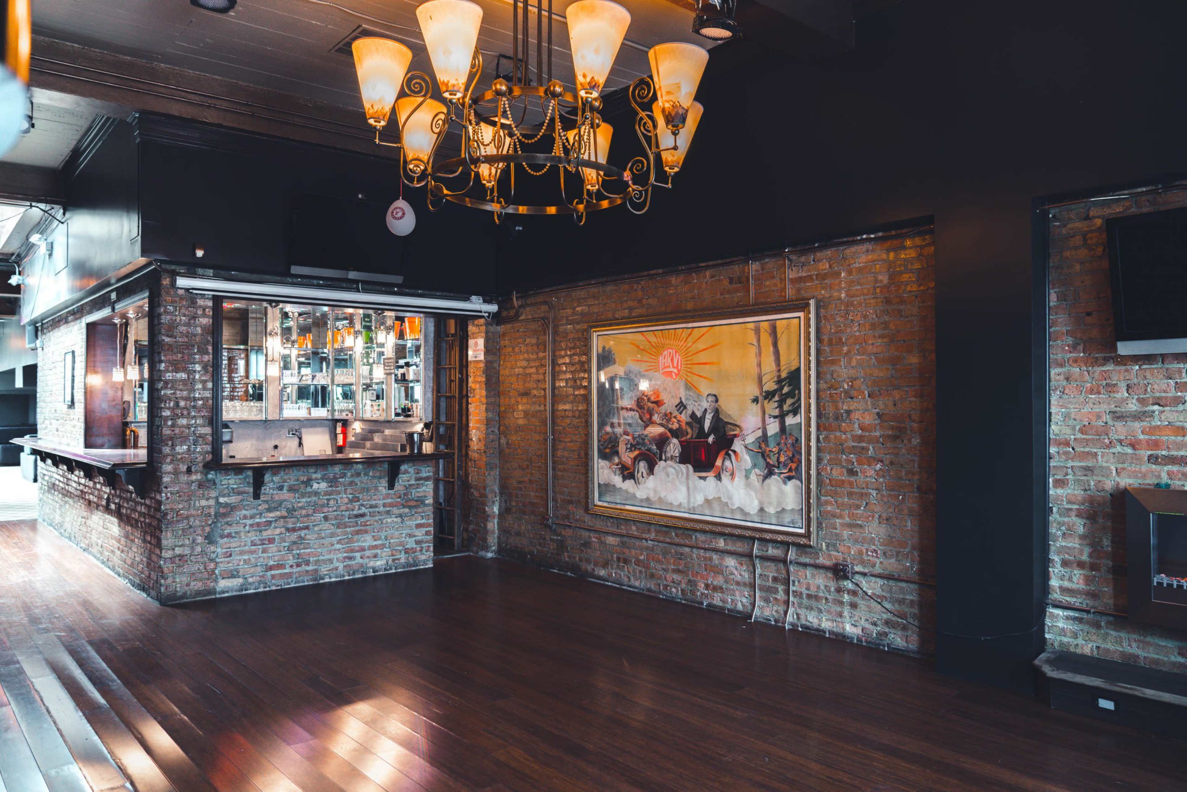 The image shows a spacious interior of a venue with exposed brick walls, wooden flooring, a large decorative painting, and a chandelier hanging from the ceiling.