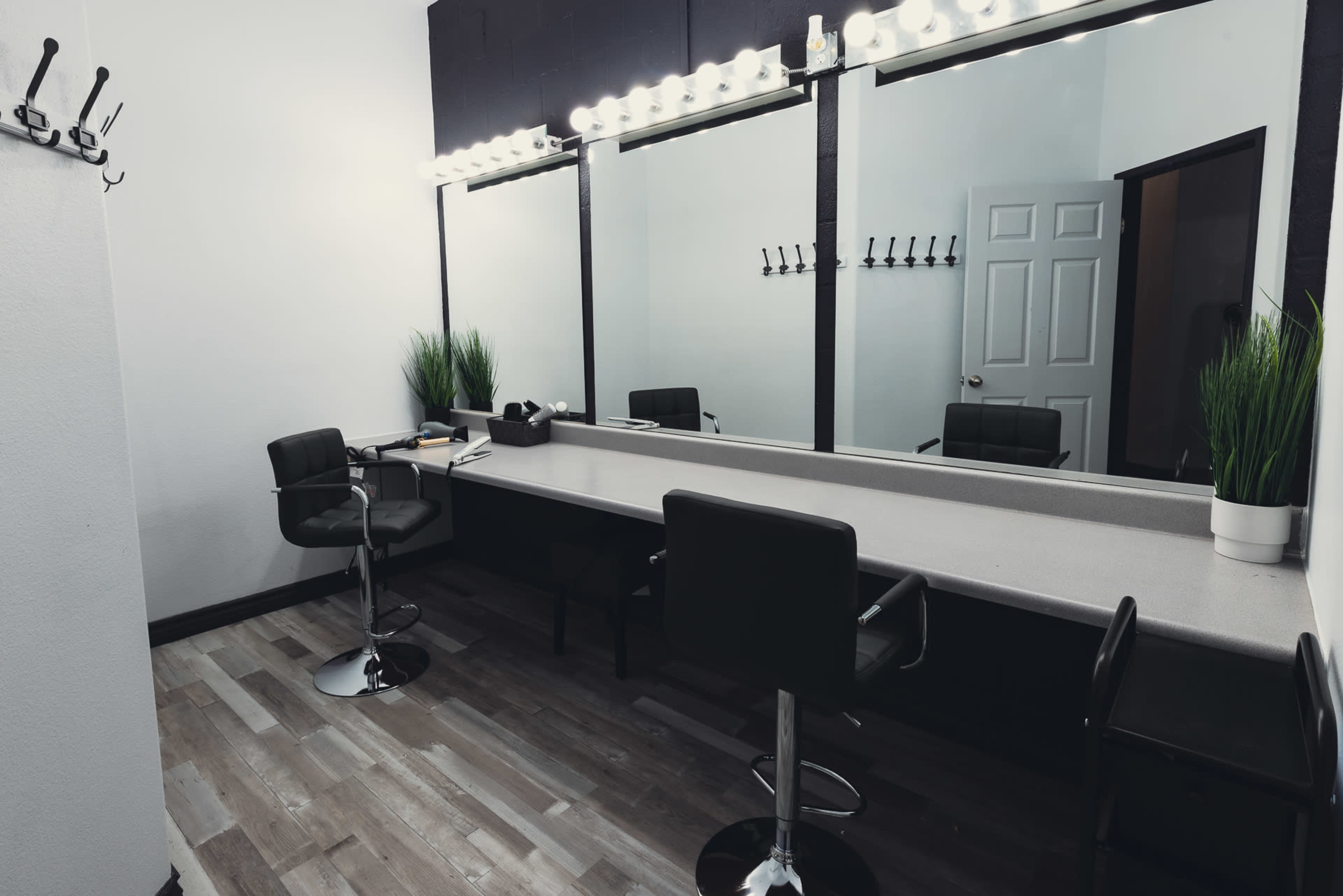 The image shows a makeup studio with a long countertop, three mirrors, black chairs, and coat hooks on the wall.