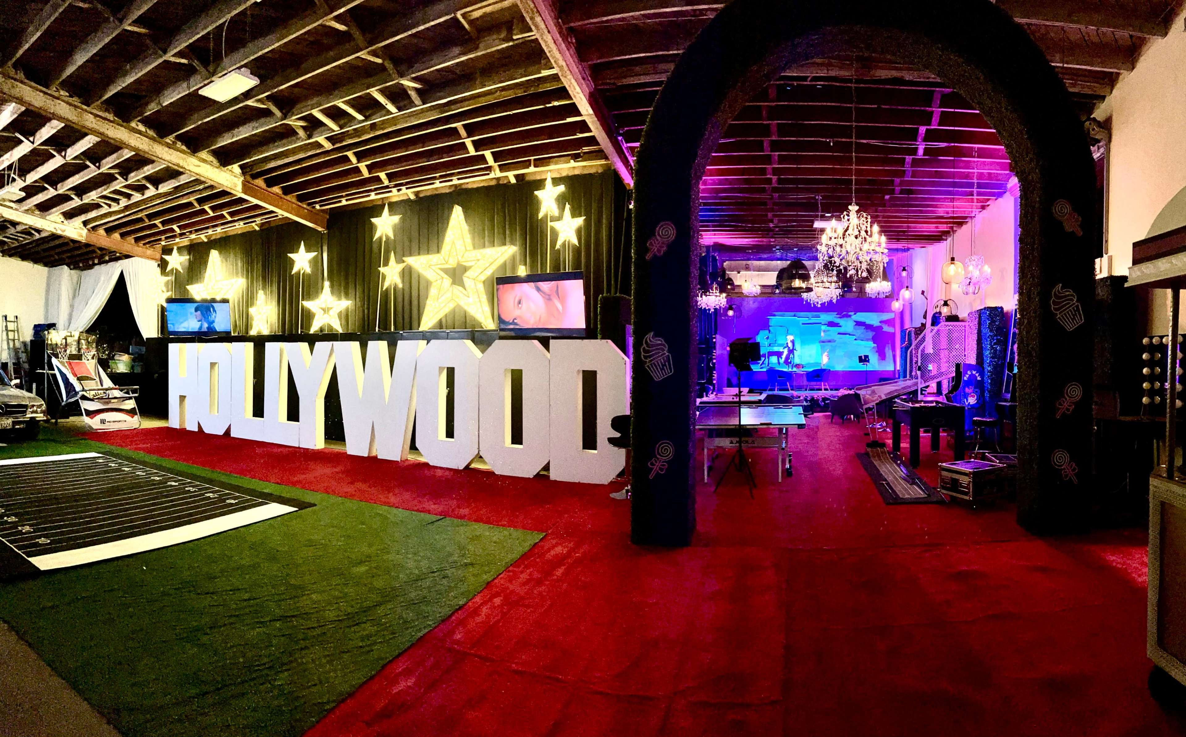 The image shows a spacious event venue decorated with a large "HOLLYWOOD" sign, star motifs, and colorful lighting, featuring a stage area with screens and a dance floor.