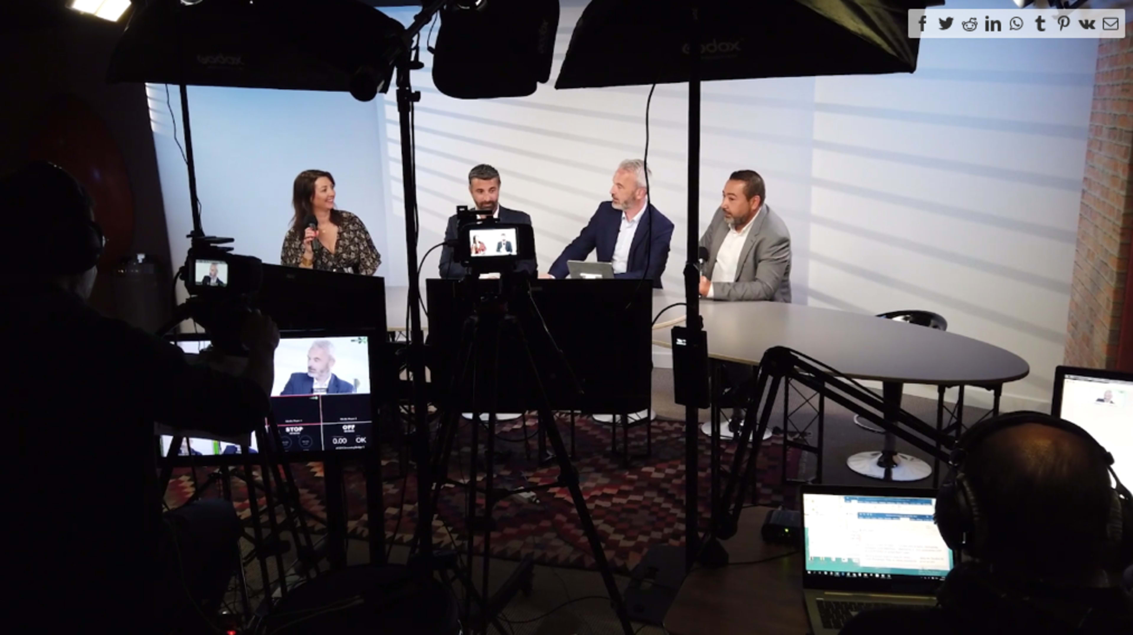 A group of four people participates in a discussion on a set equipped with cameras and lighting, while multiple screens display their conversation.