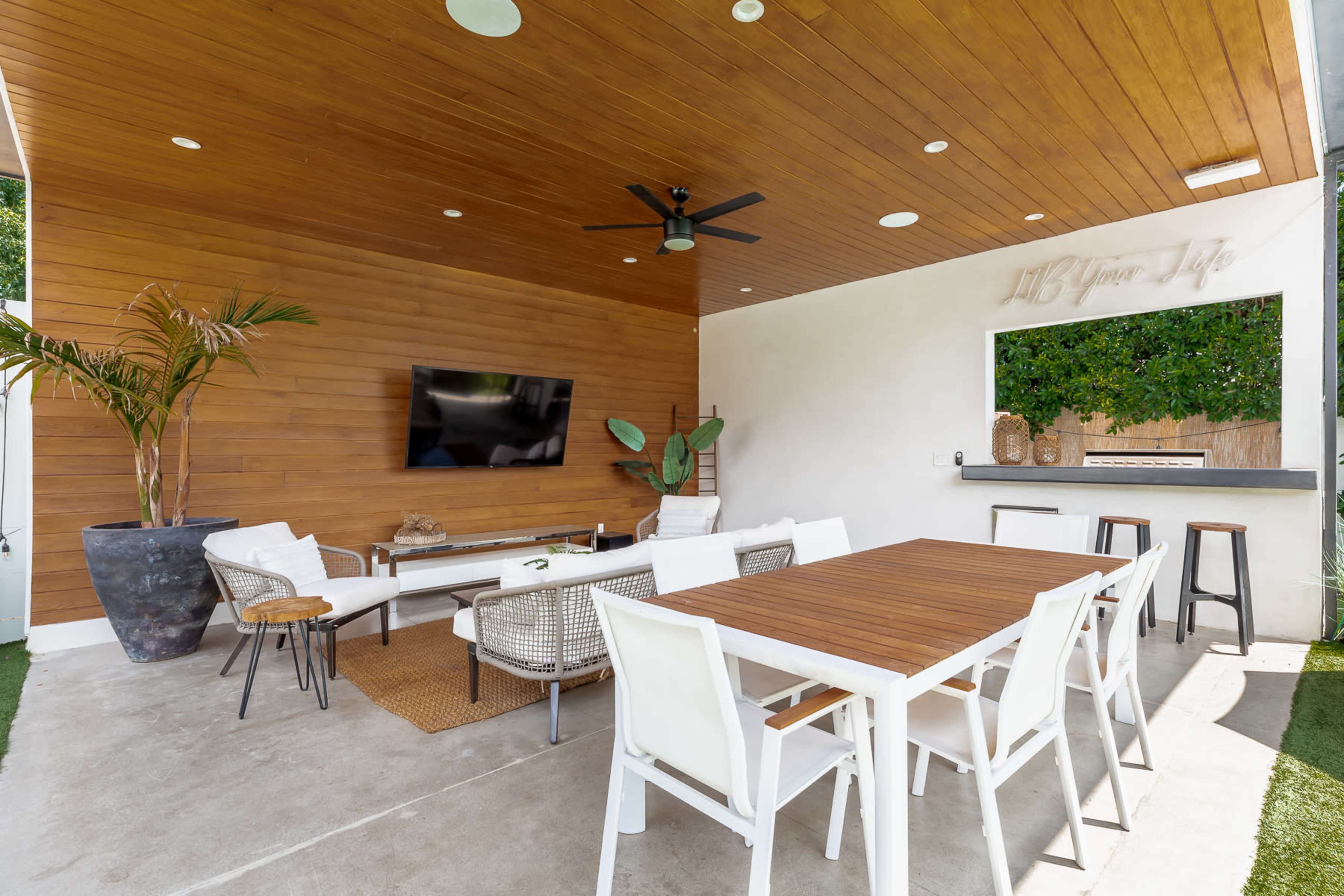 A modern outdoor patio area featuring a wooden ceiling, a dining table, a seating arrangement, a flat-screen TV, and a large planter with a plant.