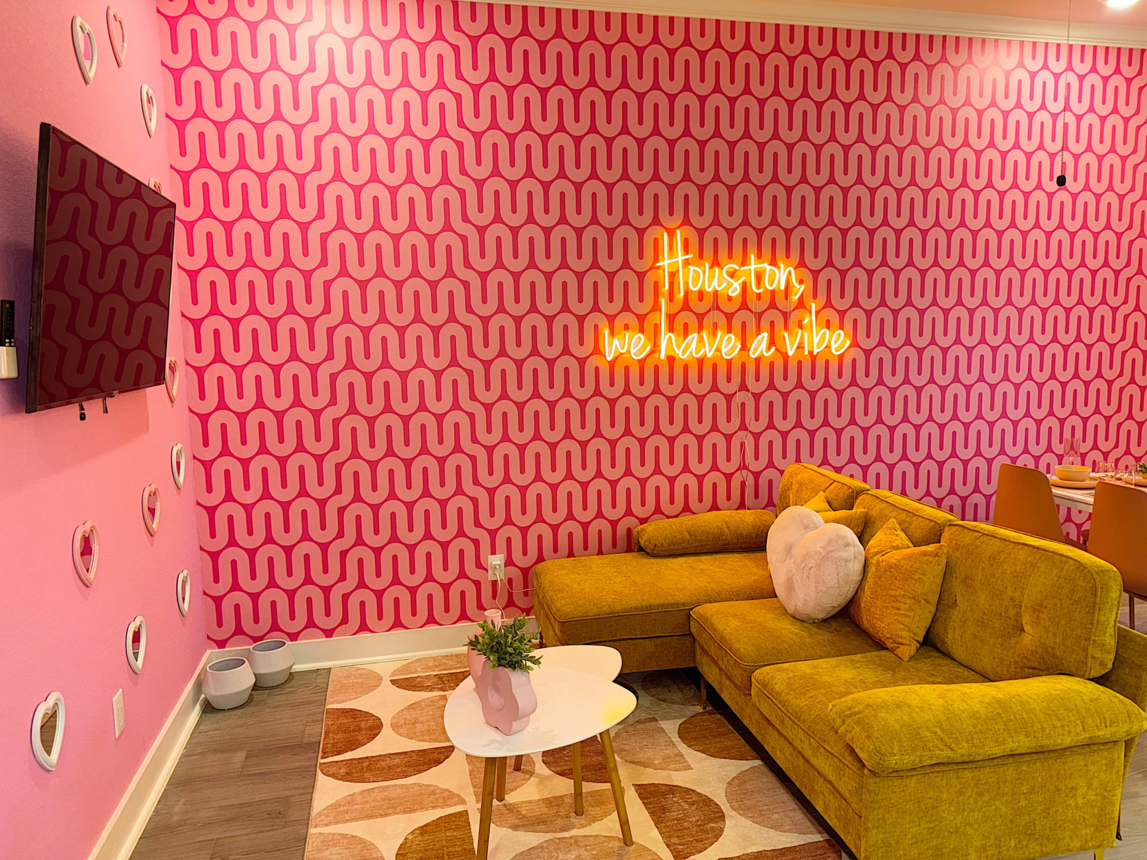 A modern living space with a pink patterned wall, a yellow sofa, an illuminated neon sign, and decorative accents.