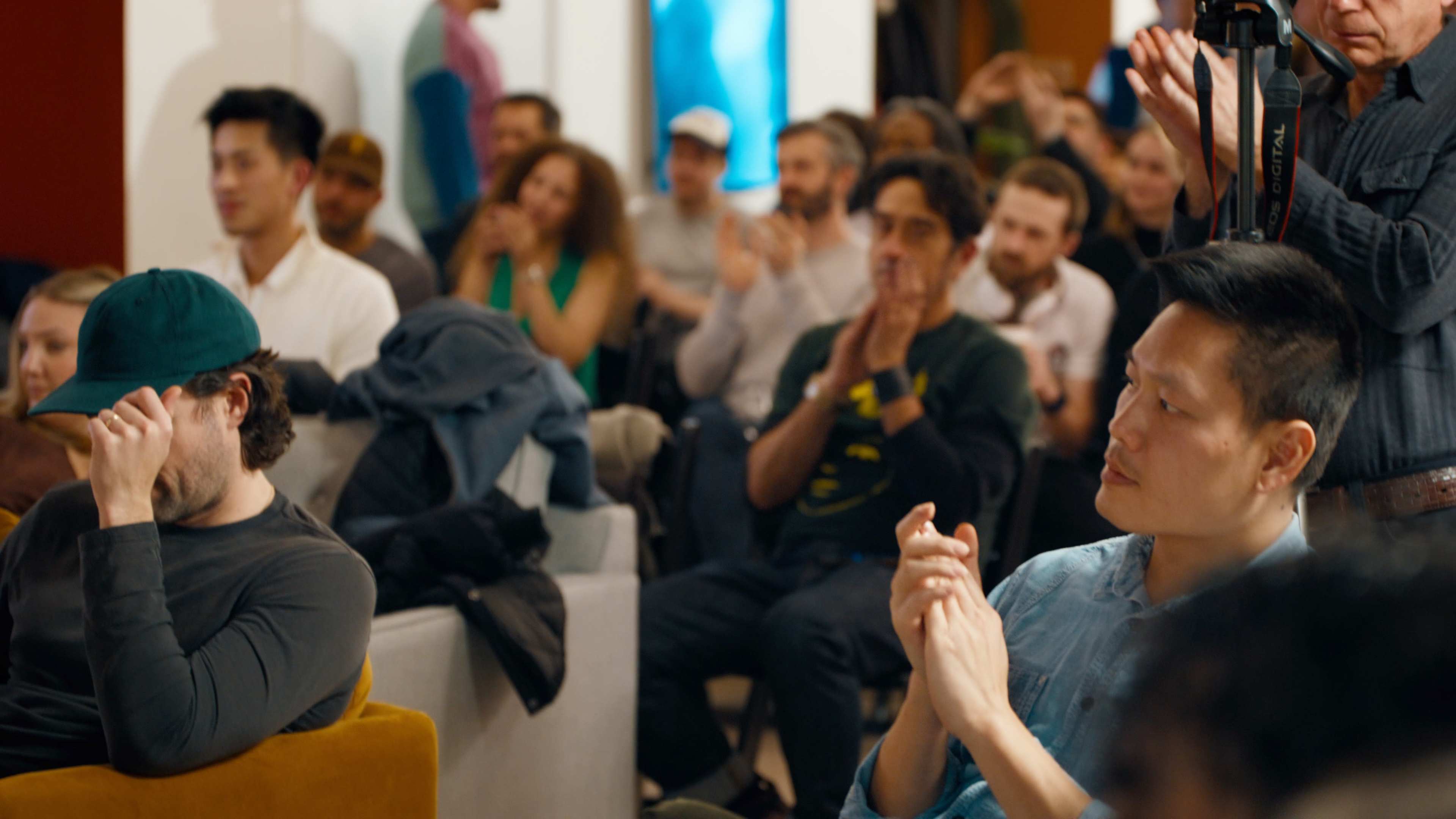 A crowd of people sits in a brightly lit room, with some attendees clapping and others looking on attentively.