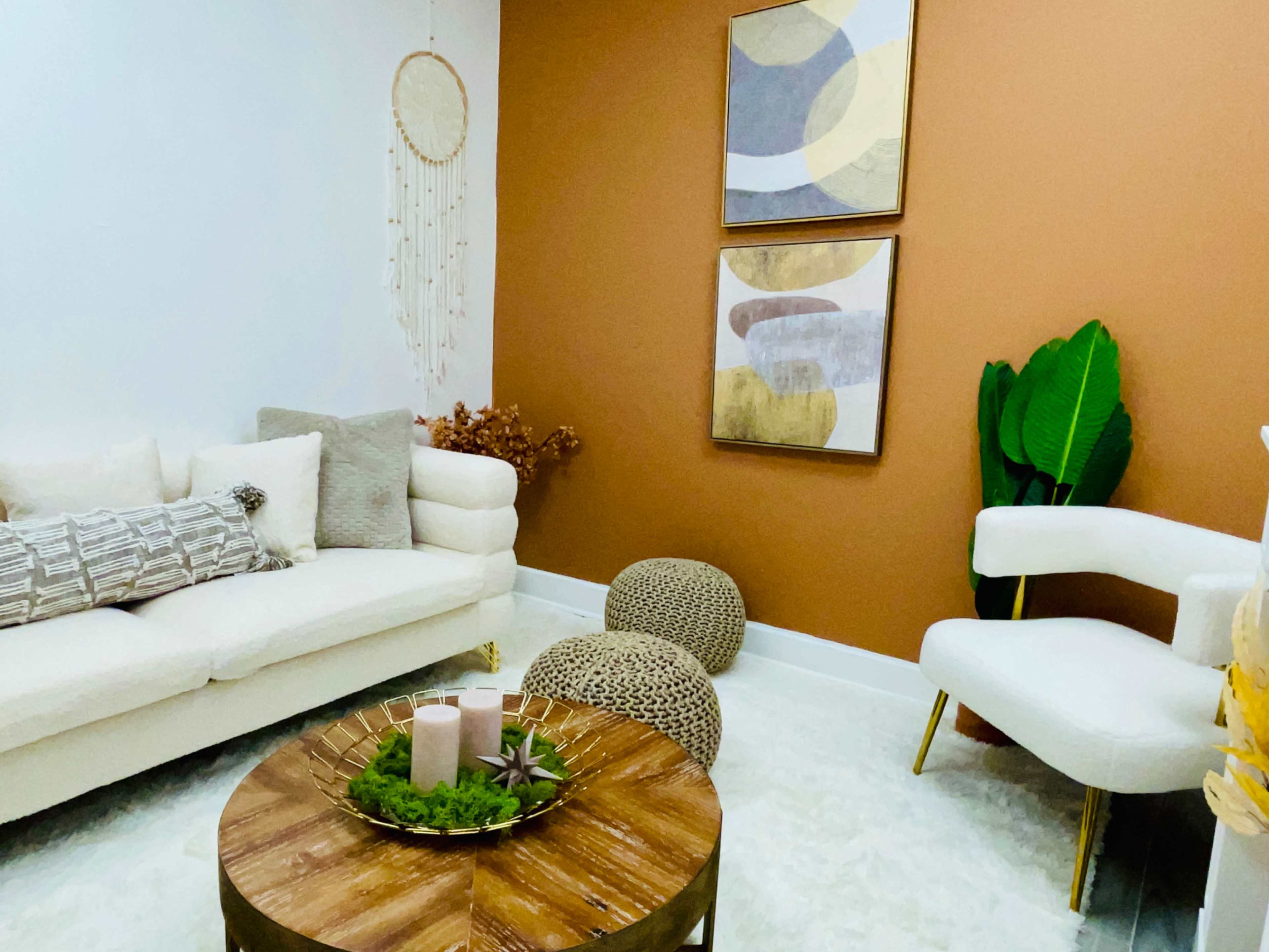 A cozy living room with a beige couch, two decorative paintings on a brown wall, and a wooden coffee table surrounded by plants and poufs.