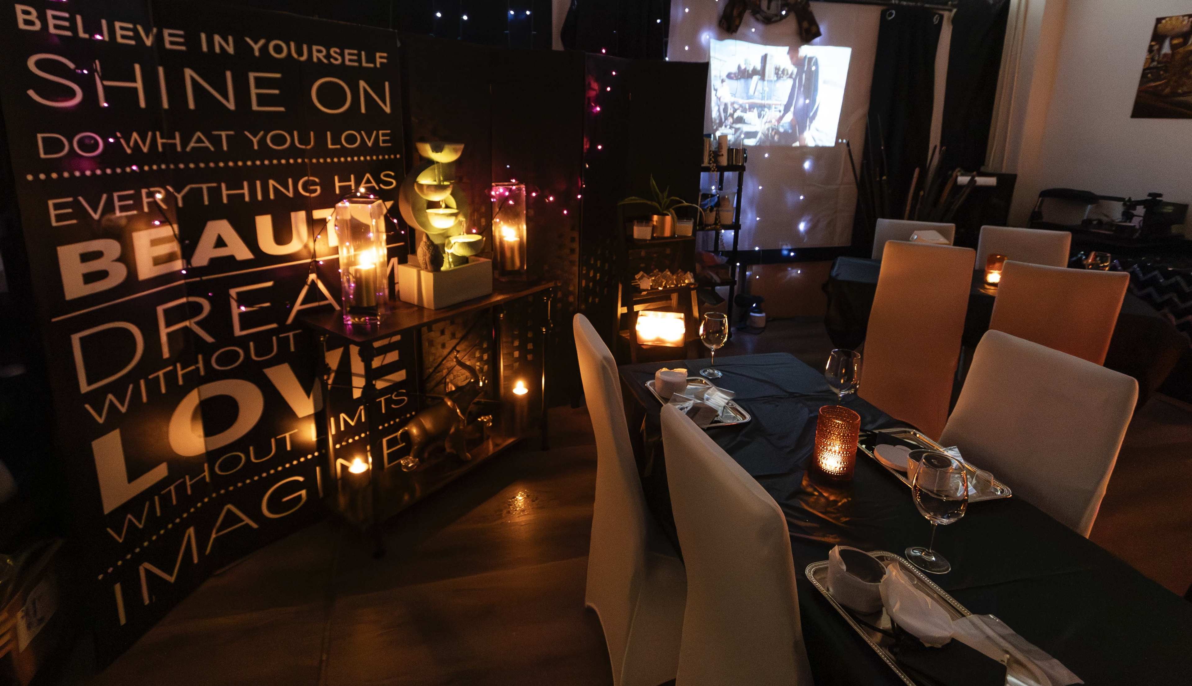 The image shows a dimly lit restaurant setting with tables arranged for dining, featuring decorative candles, a motivational wall display, and a projector displaying an image on a screen.