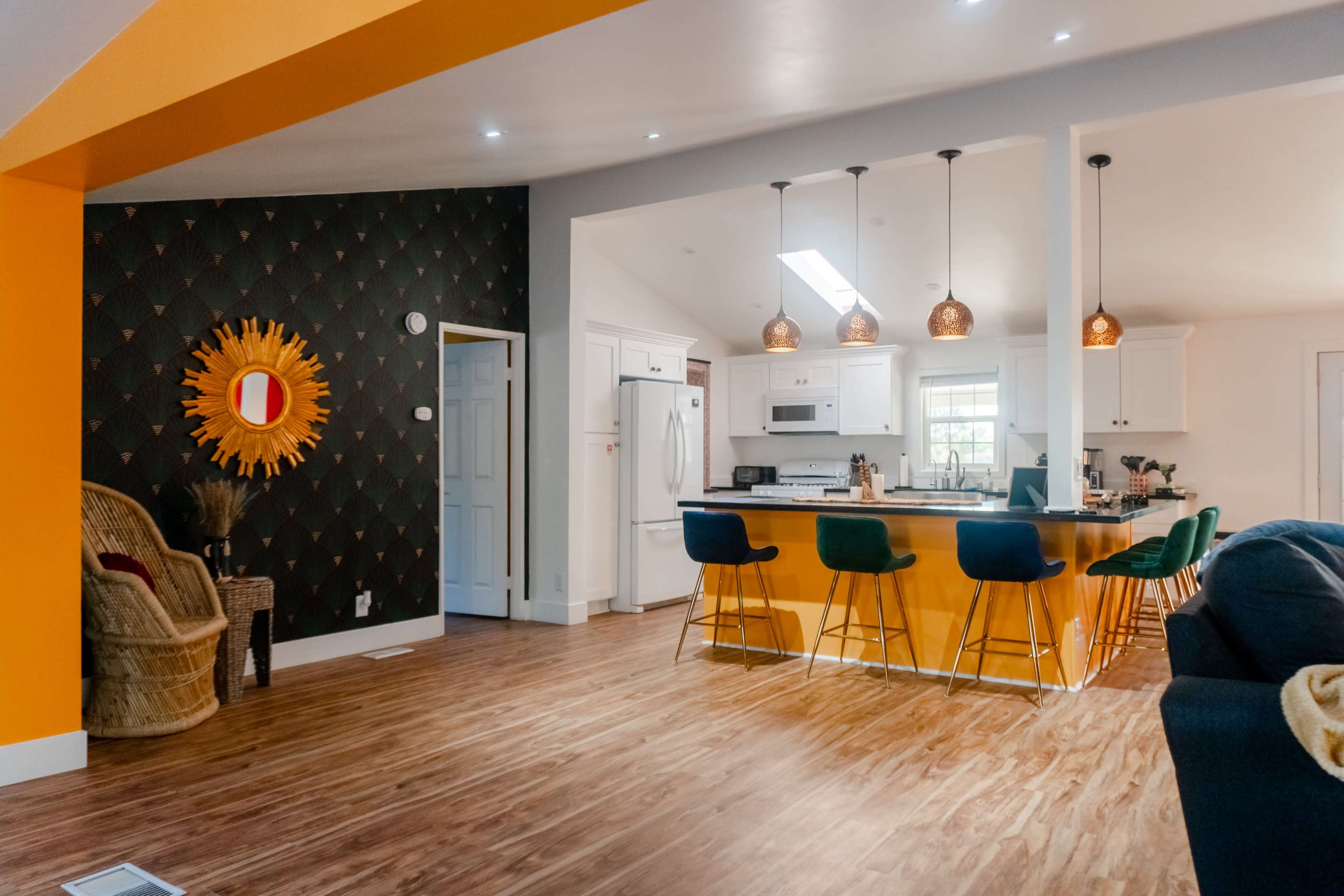 A modern kitchen area features a yellow accent wall, pendant lights, and a bar with colorful stools, alongside a cozy seating area with a wicker chair.