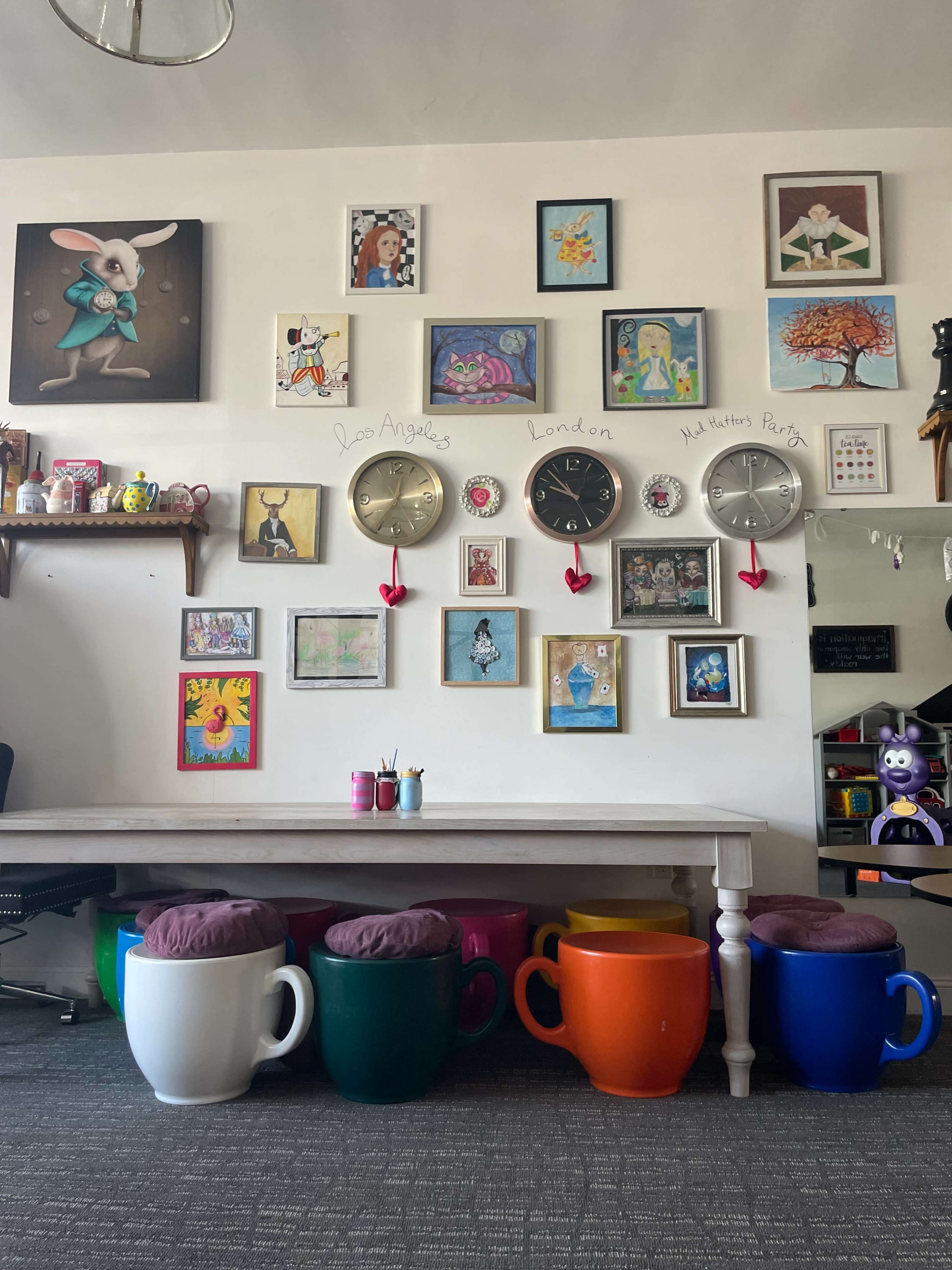 The image shows a brightly decorated wall filled with various framed artworks and clocks, alongside a large table with colorful mug-shaped stools.