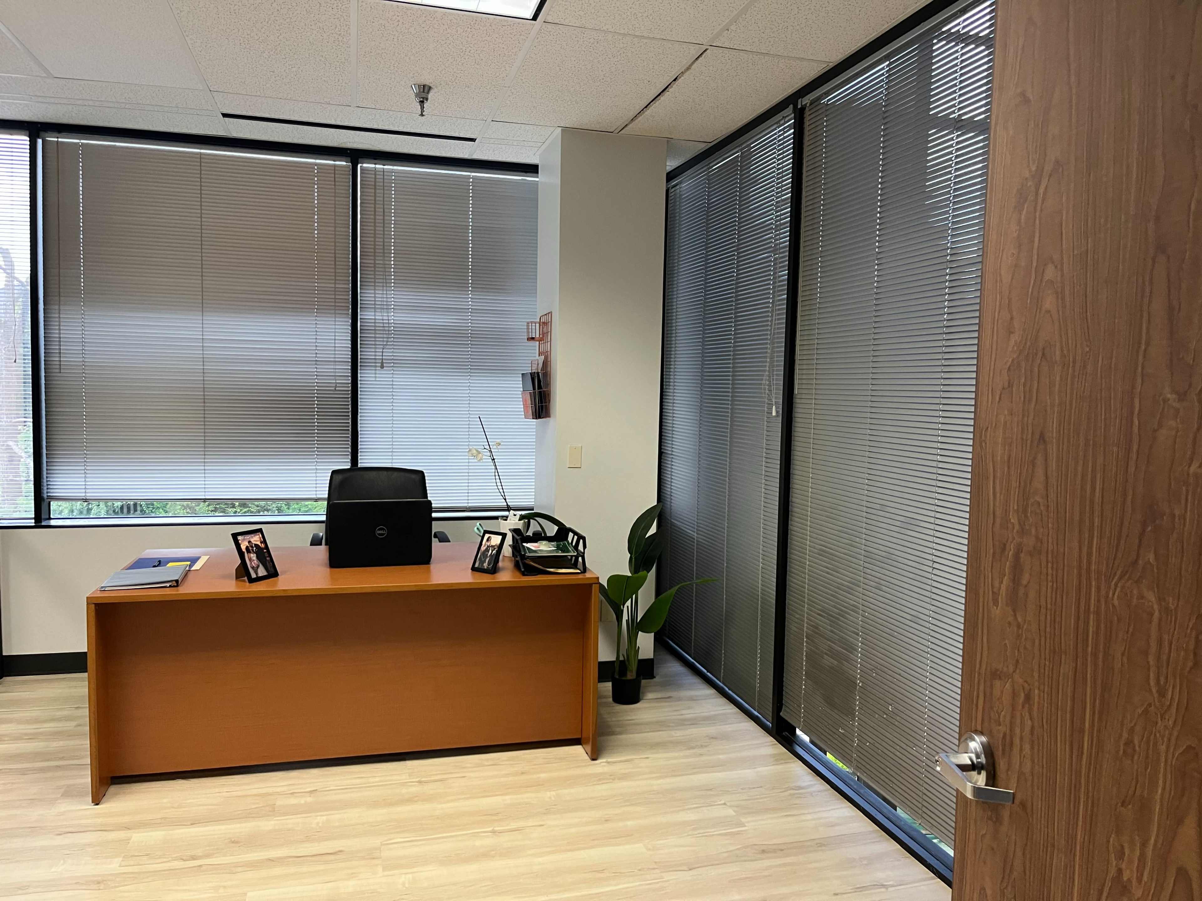 The image shows a modern office with a wooden desk, a chair, and two framed photos on the desk, alongside a potted plant and large windows with blinds partially drawn.