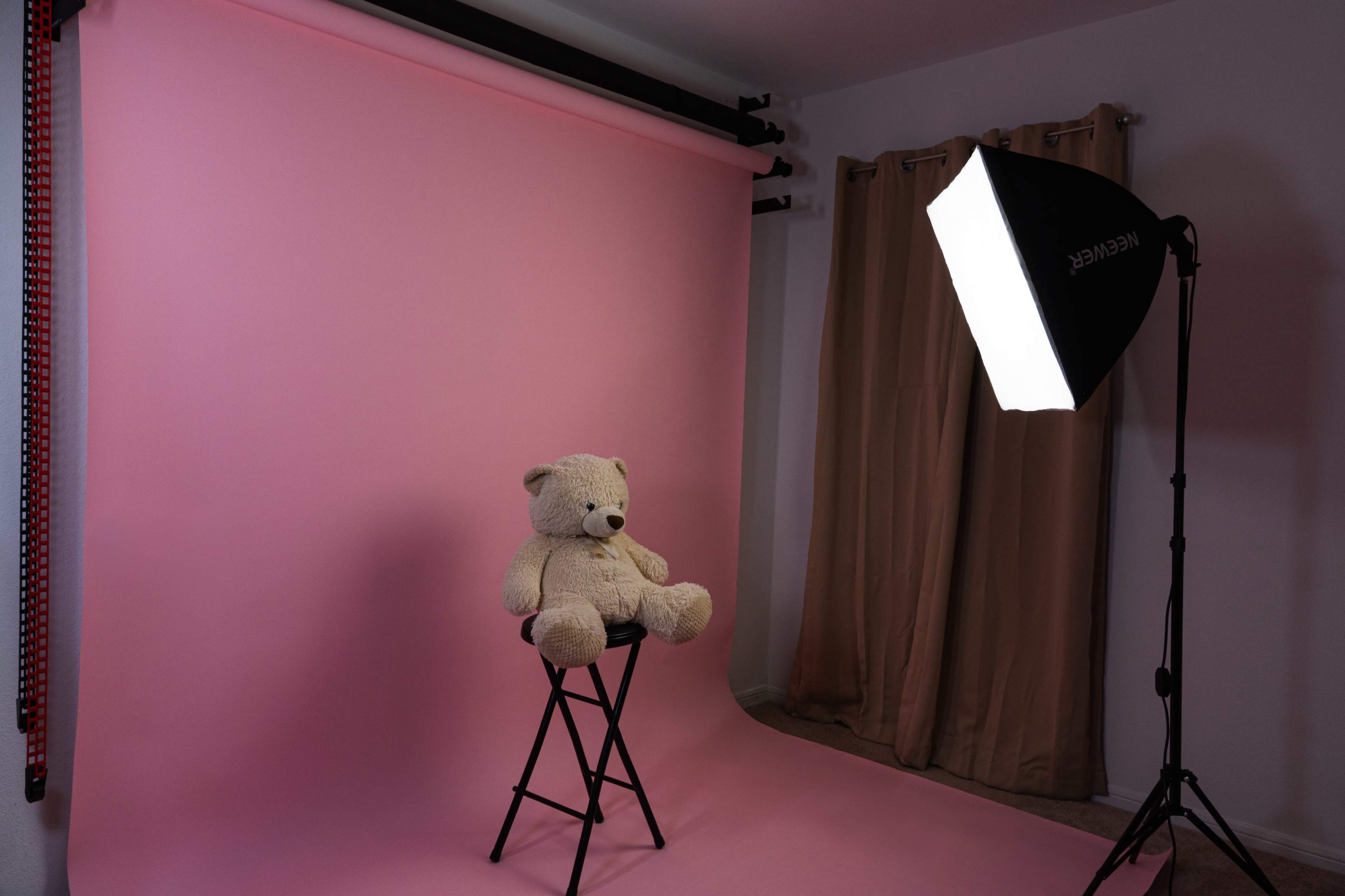 A teddy bear sits on a stool in a photography studio with a pink backdrop and a softbox light.