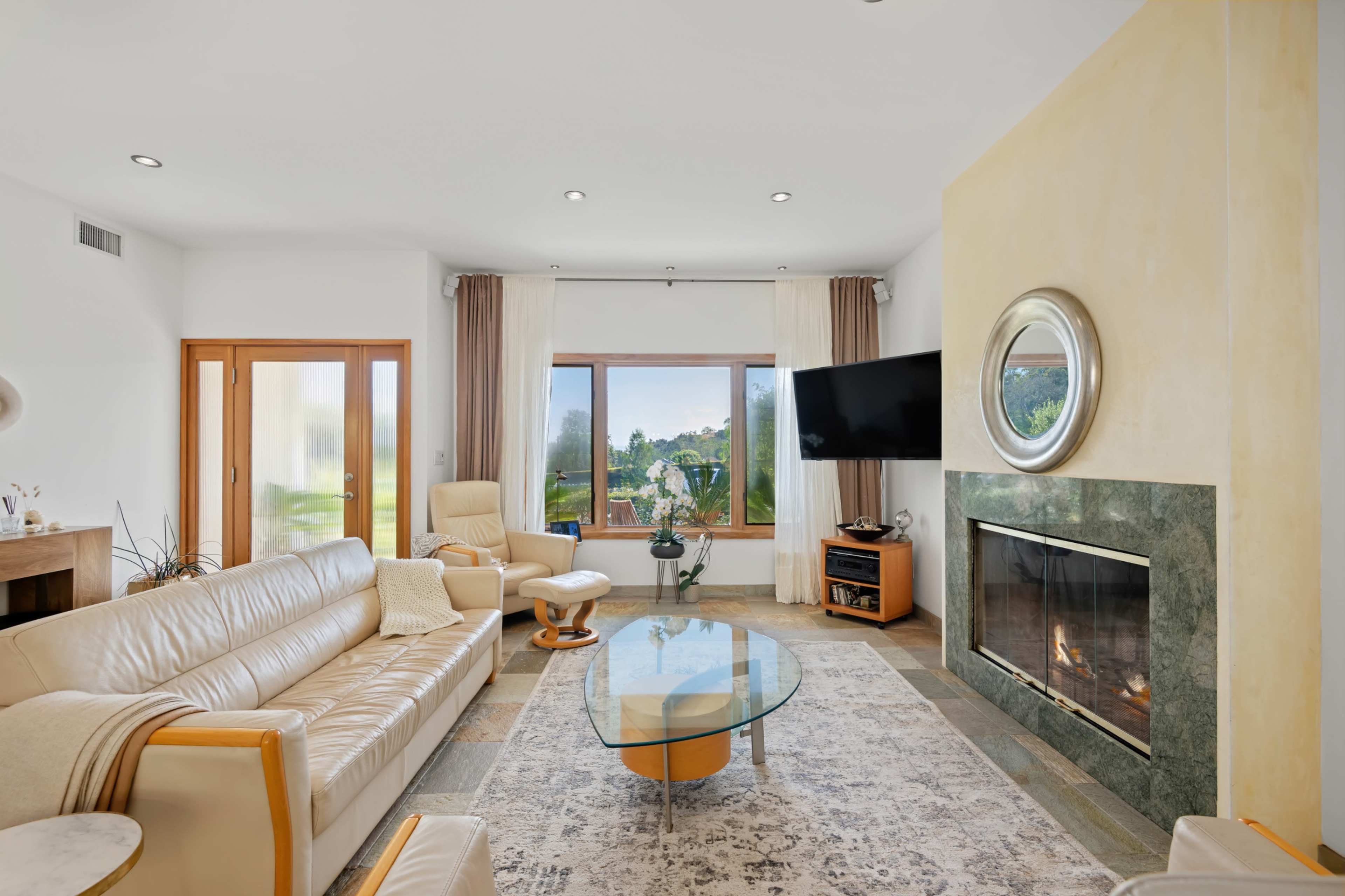 The living room features a beige leather sofa, a glass coffee table, a modern fireplace, and large windows providing natural light and a view of the outdoors.