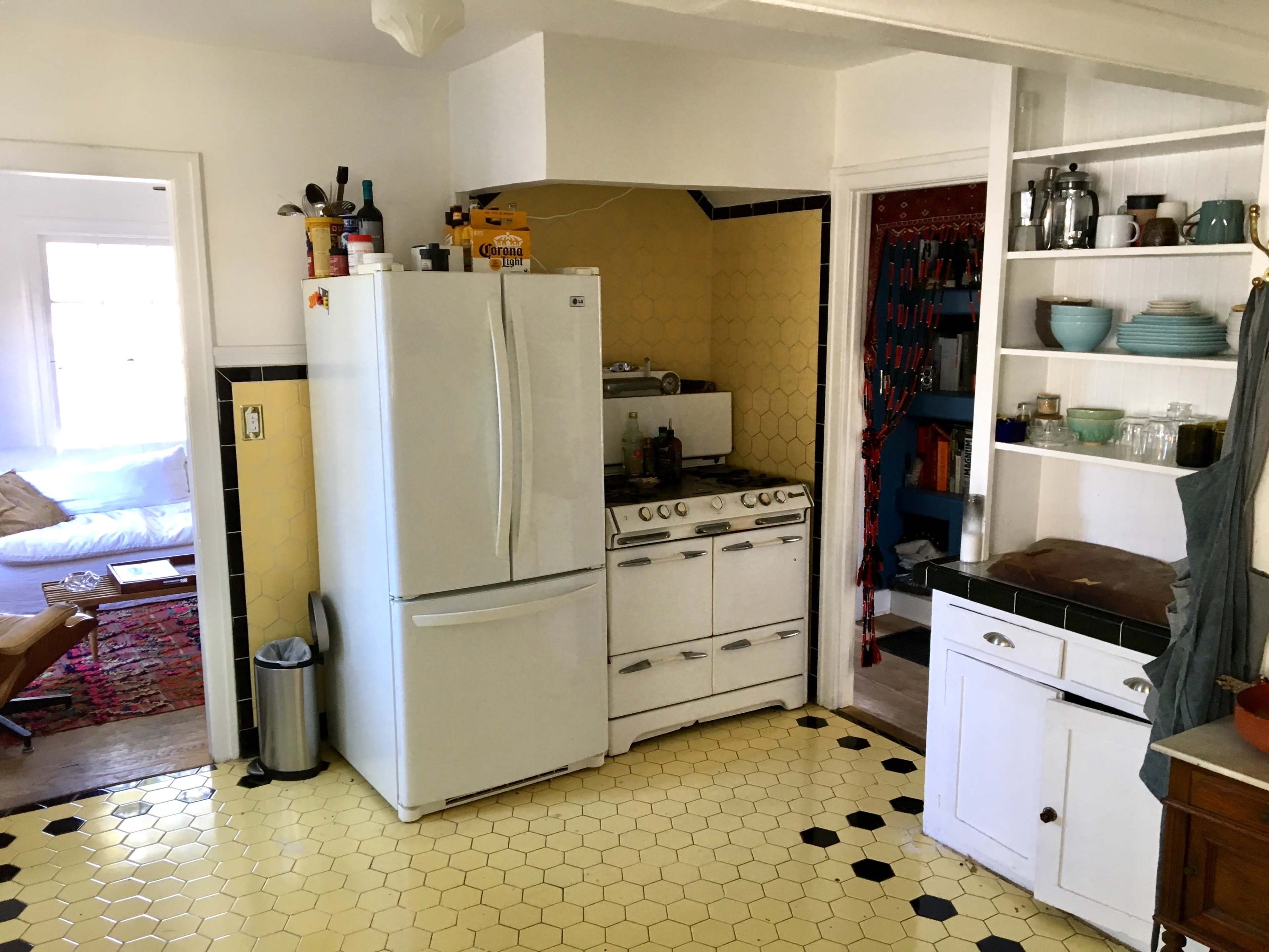 The image shows a kitchen with a white refrigerator, a stove, yellow hexagonal tiles on the floor, and shelves displaying dishes and glassware.